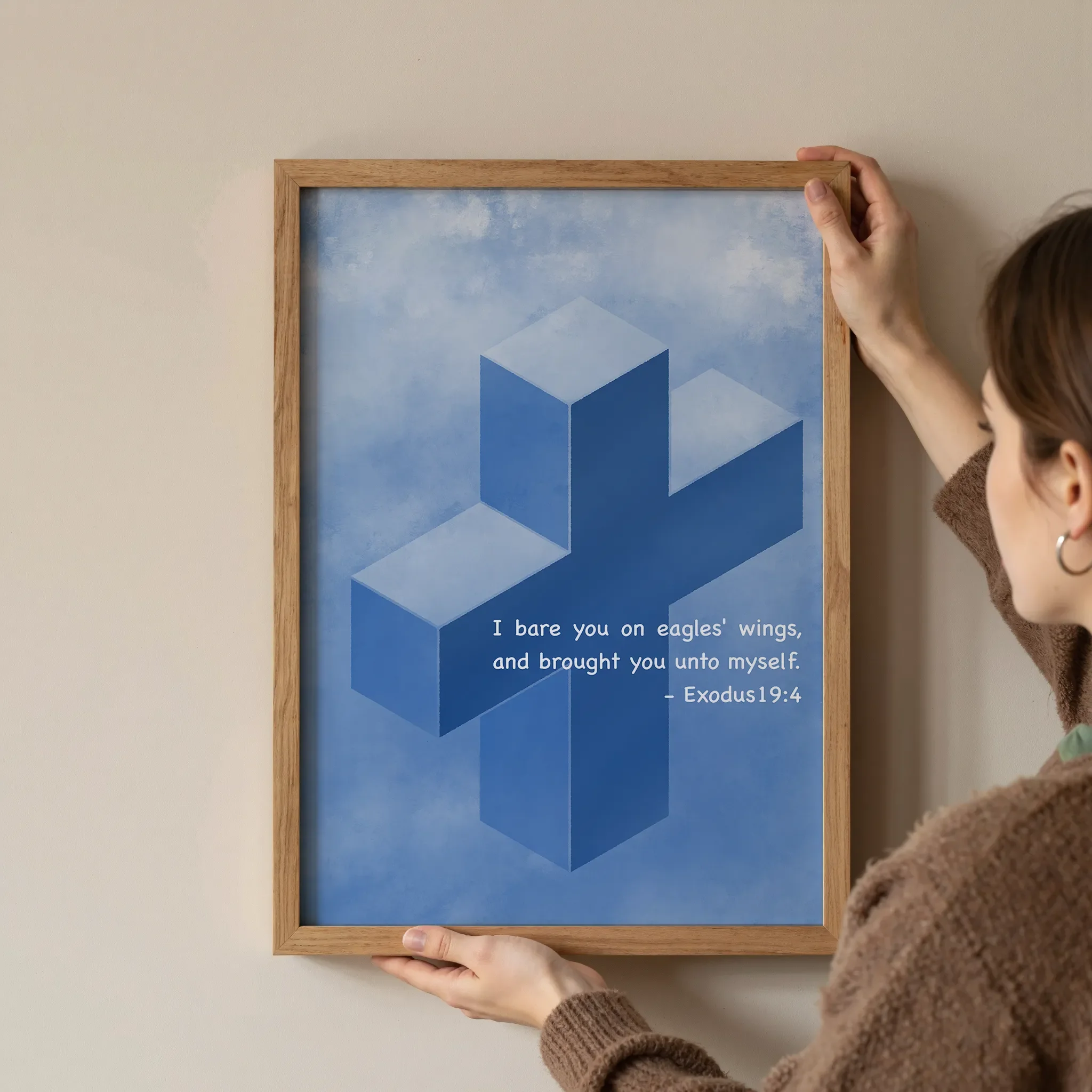 A Scripture wall art featuring a cross in a simple wooden frame is held at the corners by a young woman against a neutral wall.