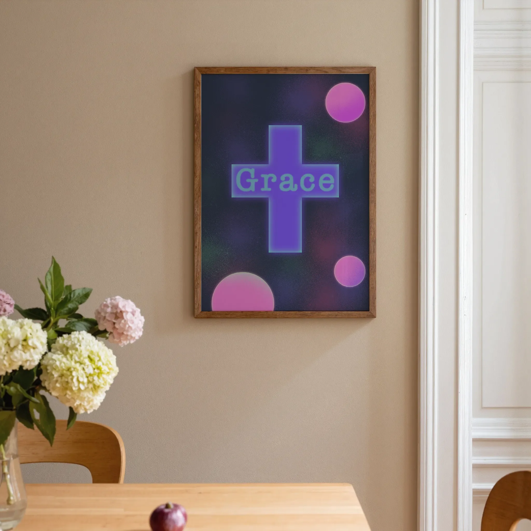 A colorful geometric cross print hangs on a warm neutral wall beside a white door in a home interior. In front is a warm wooden table with pink and pale yellow flowers. The room is softly lit.