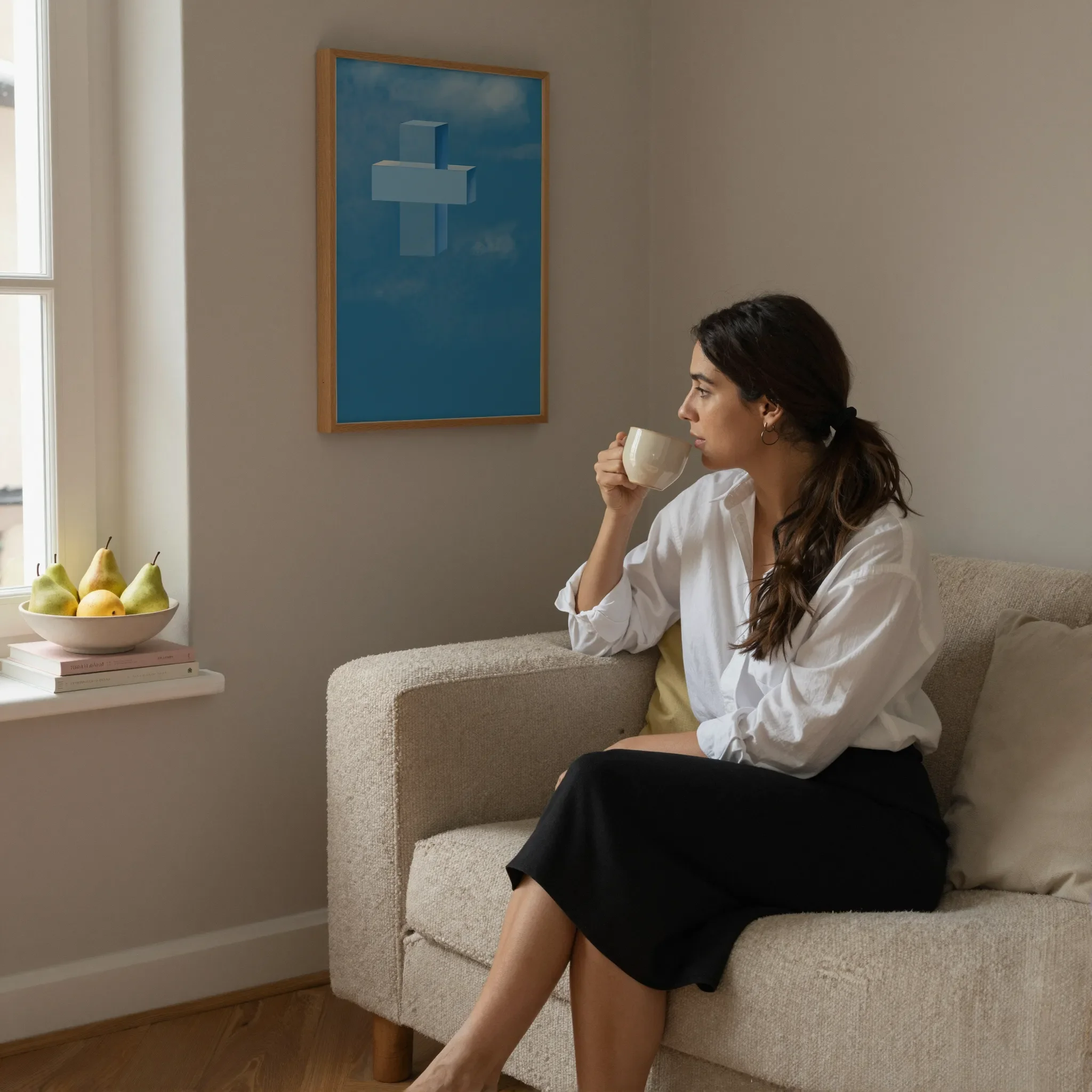 A cross print hangs on a neutral wall in a cozy living room. A woman sits on a sofa holding a ceramic cup and looking out the window. Pears and books rest on the windowsill.