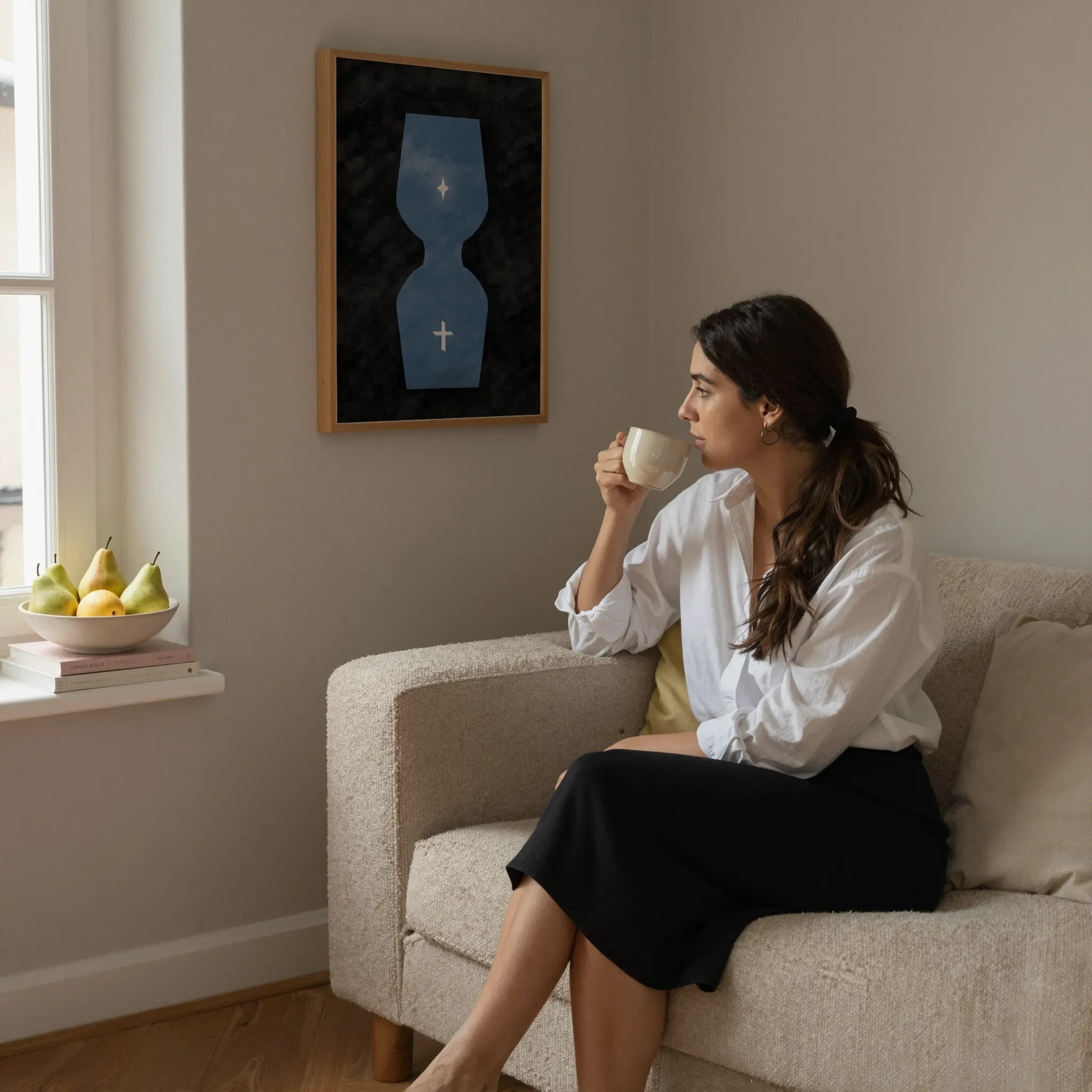 A surreal hourglass Christian print hangs on a neutral wall in a cozy living room. A woman sits on a sofa holding a ceramic cup and looking out the window. Pears and books rest on the windowsill.