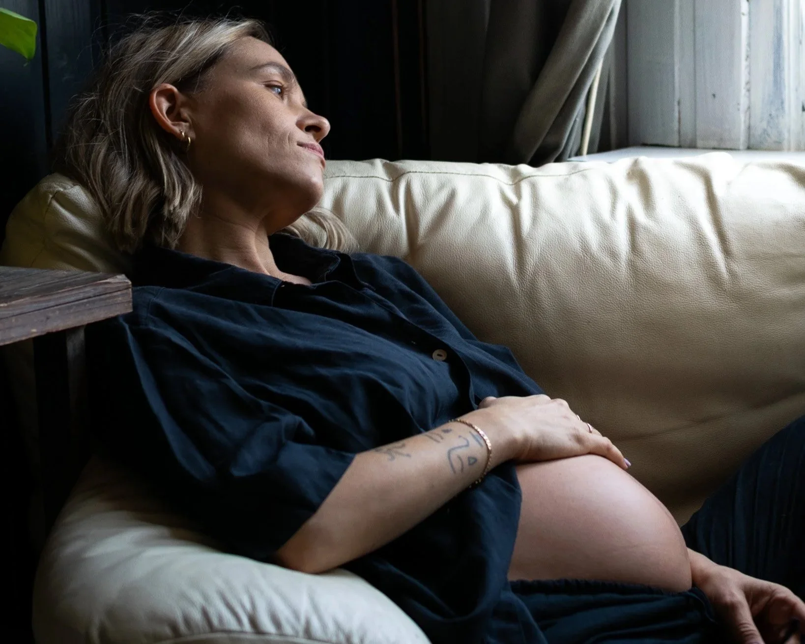 Vrouw in blauwe doorzichtige lingerie met bloemenpatroon, zittend op haar knieën op een vloerkleed, met haar handen achter haar hoofd in een kamer met een vintage bank en een plant op de achtergrond.