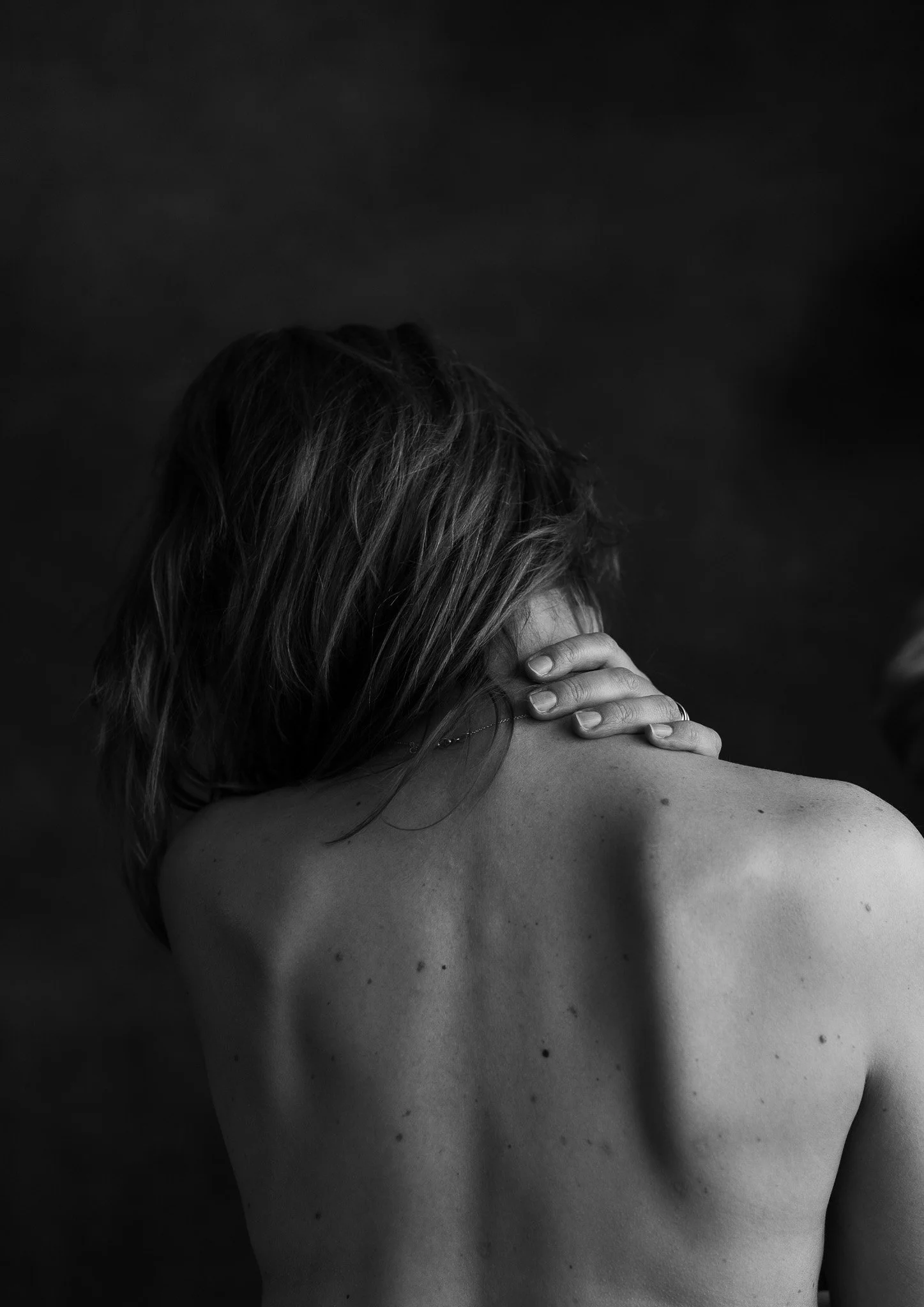 Zwart-wit foto van een vrouw die haar nek vasthoudt, haar rug en schouder zichtbaar, warrig haar dat haar gezicht bedekt.