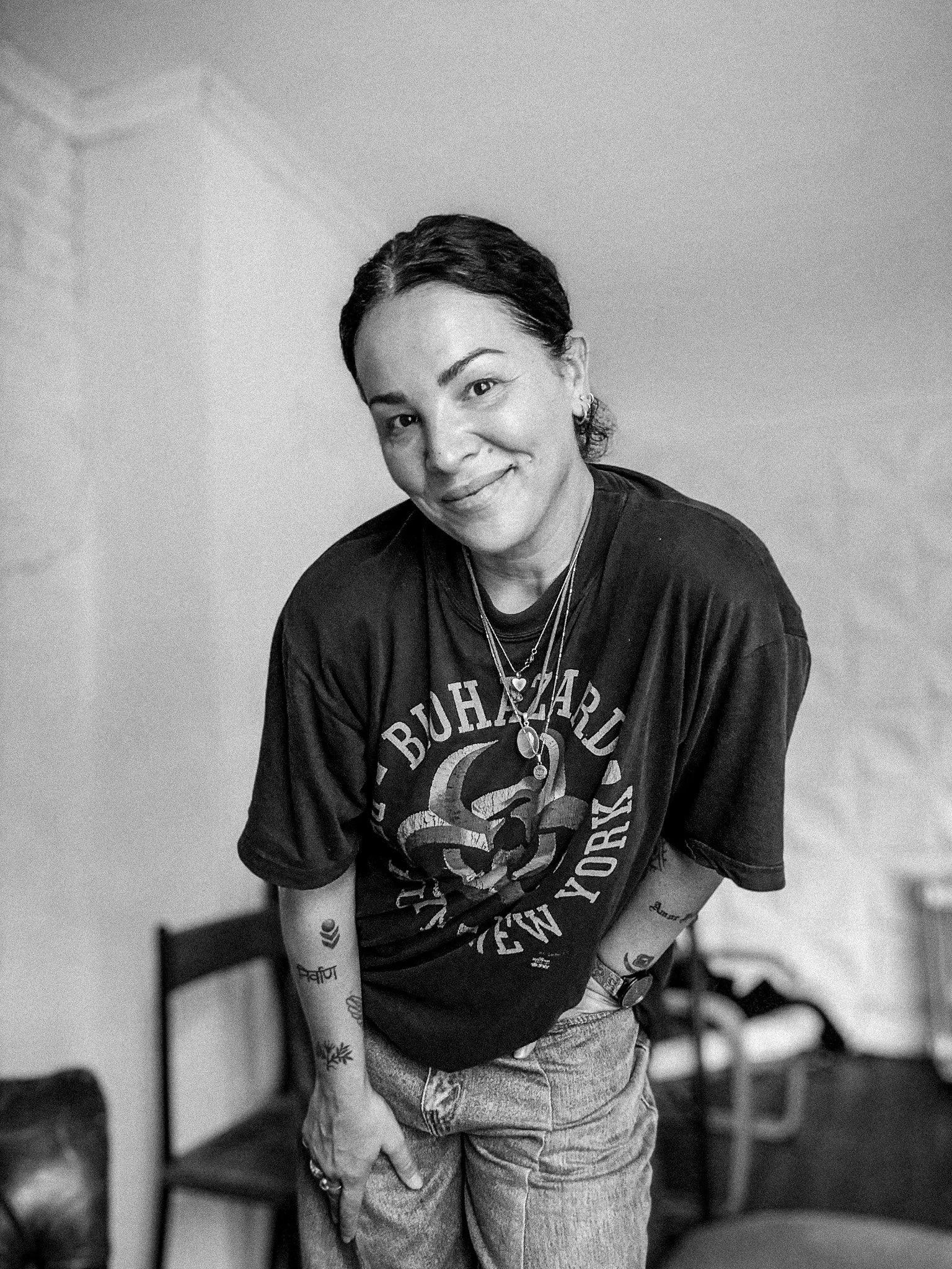 Een vrouw met kort donker haar die glimlacht, gekleed in een zwart T-shirt en spijkerbroek, met meerdere sieraden en tattoos, zittend tegen een witte muur.