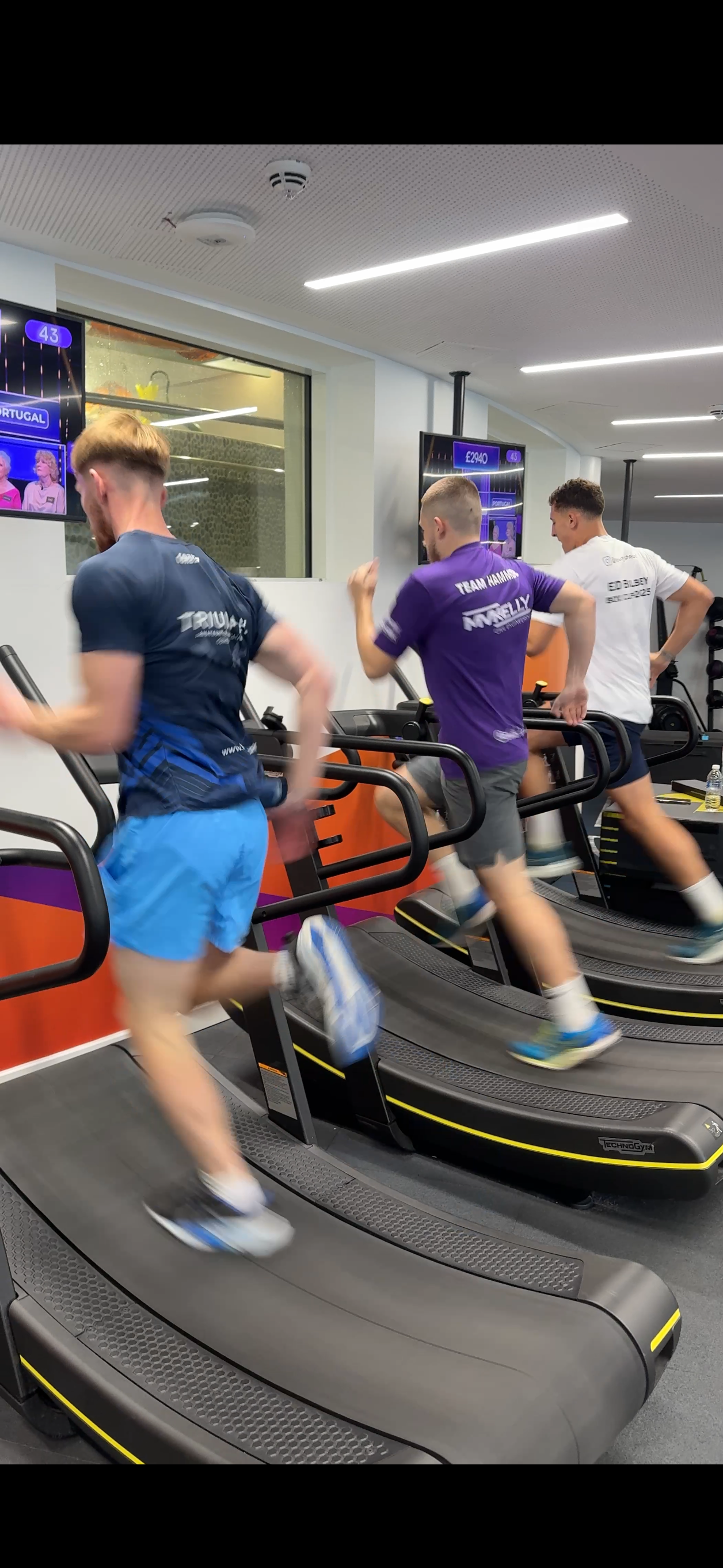 Three men running on three treadmills in a gym, with mirrors and digital screens in the background.