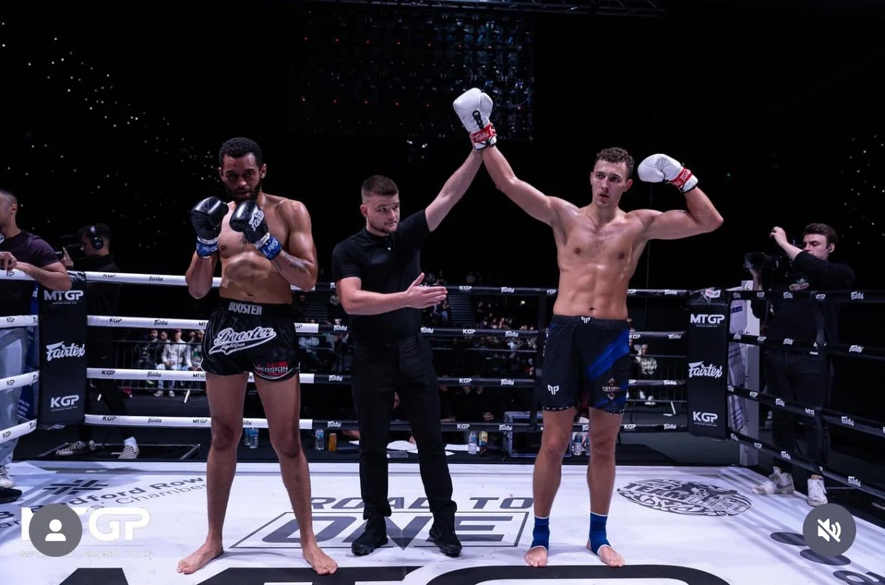 Two male fighters in a boxing ring, one with black shorts and the other with blue shorts, are celebrating after a match. The man in black shorts has a serious expression, while the man in blue shorts is raising his arm in victory. A referee is standing between them, raising the arm of the winner. There are cameras and other people around the ring.