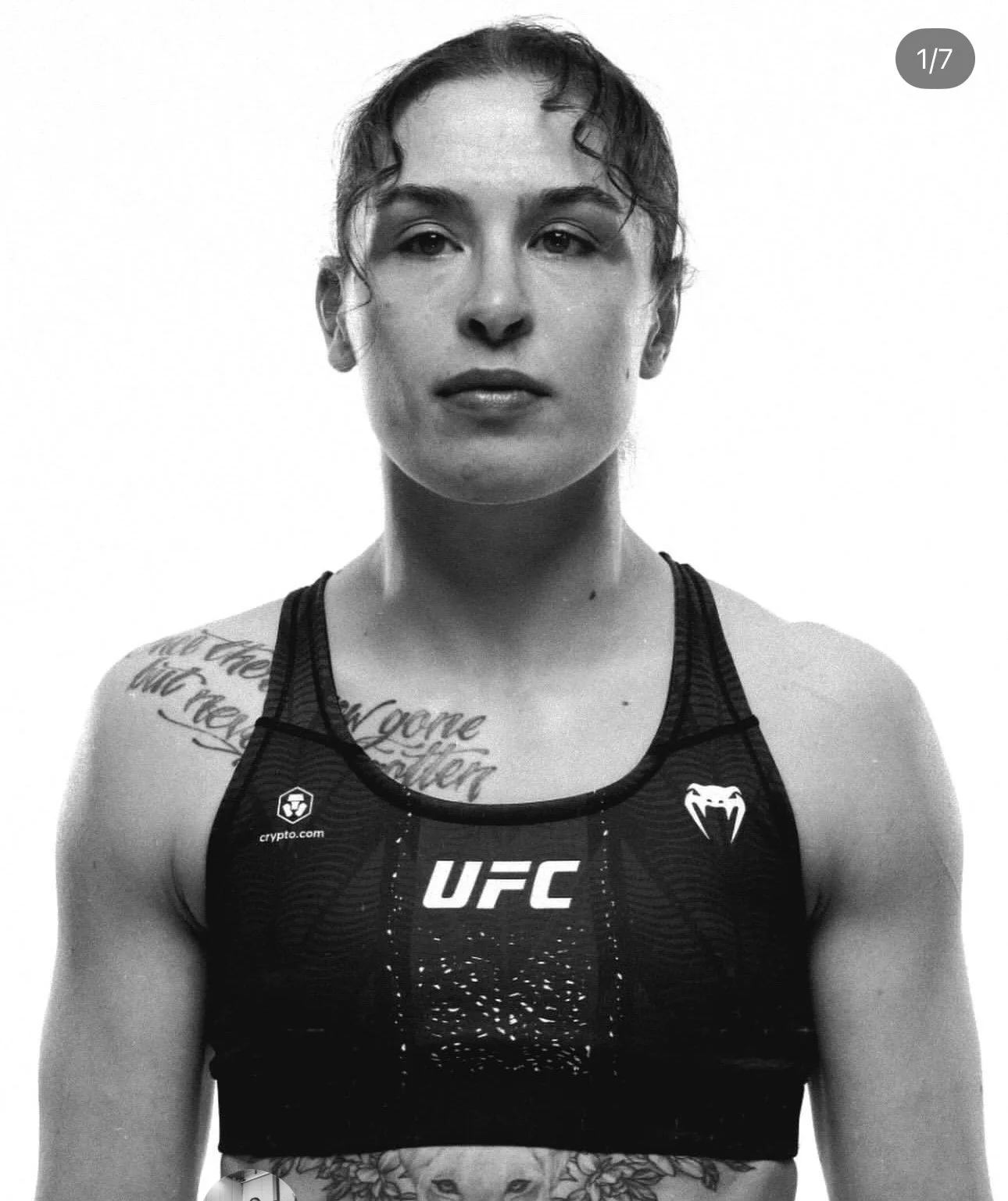 Black and white photo of a woman with short wavy hair wearing a sports bra with UFC and Venom logos. She has a tattoo on her right shoulder and chest and is looking directly at the camera.