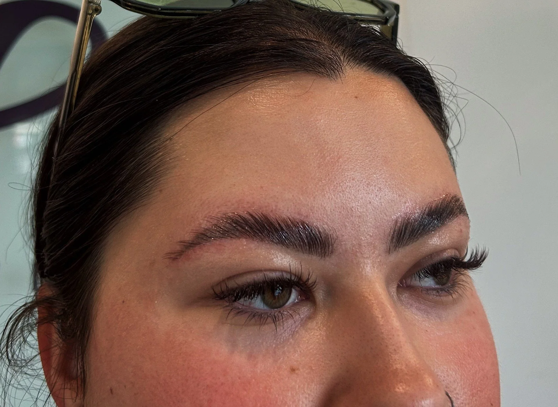 Close-up of a person's face focusing on well-groomed eyebrows and eye makeup, with glasses resting on top of their head.