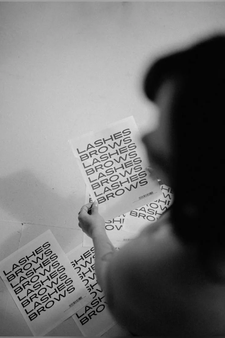 Person holding multiple sheets of paper with the words 'LASHES' and 'BROWS' printed repeatedly in bold uppercase letters.
