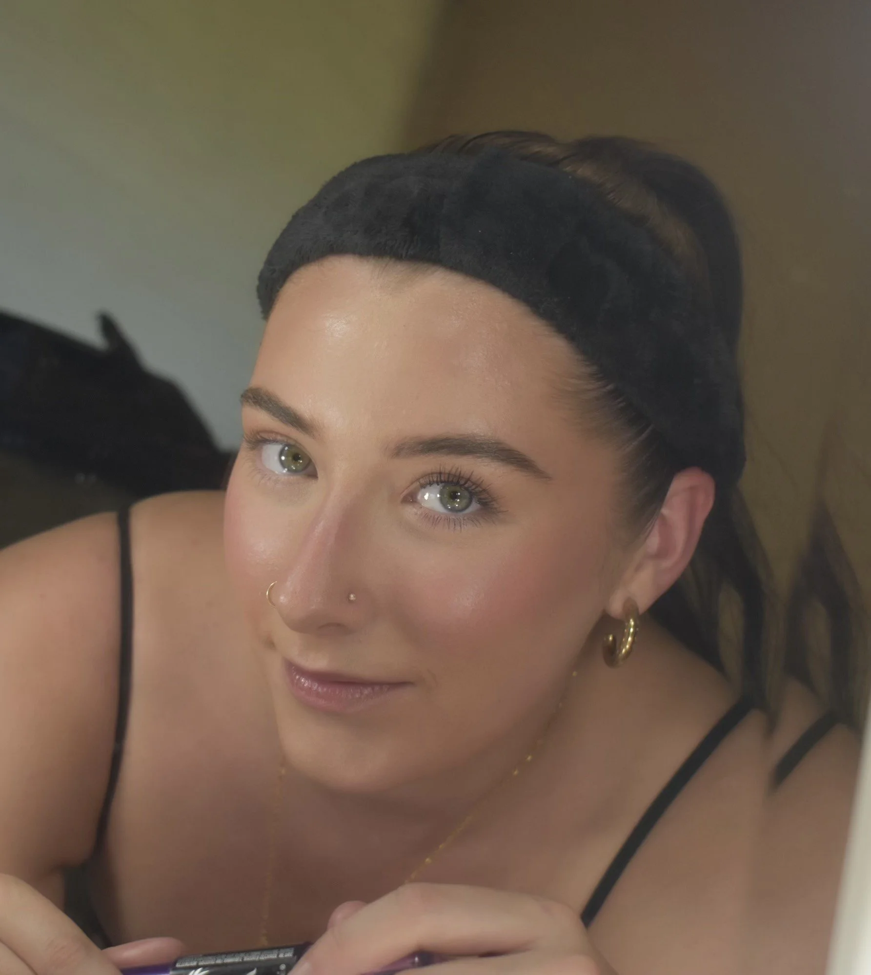 A young woman with light skin, blue eyes, and dark hair, wearing a black headband and gold hoop earrings, looking at the camera with a slight smile.