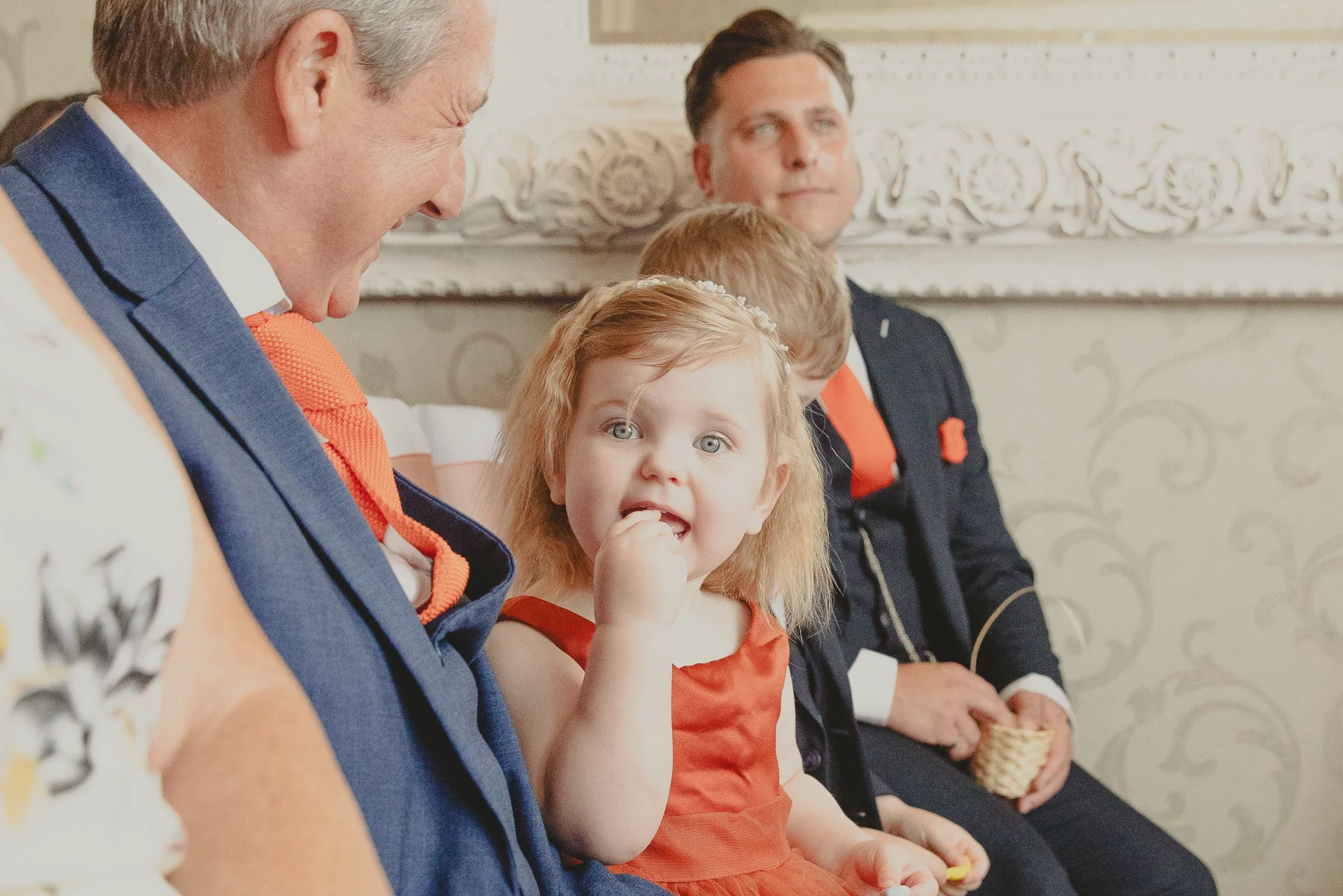 A young girl with red hair and blue eyes, wearing an orange dress, sitting between two men in suits at a formal event, as she looks directly at the camera with her finger near her mouth.