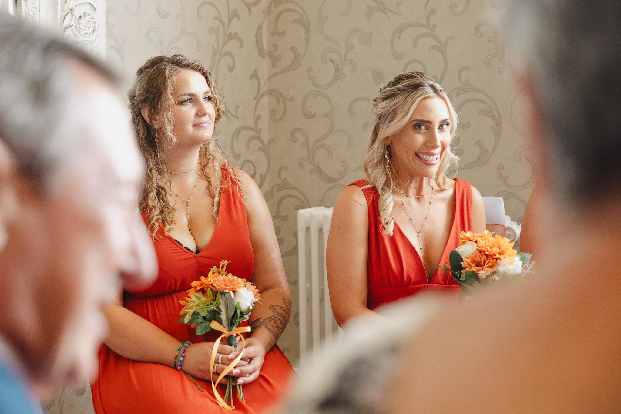 Two women in matching red dresses holding bouquets, sitting in a room with patterned wallpaper, smiling and listening to an event.