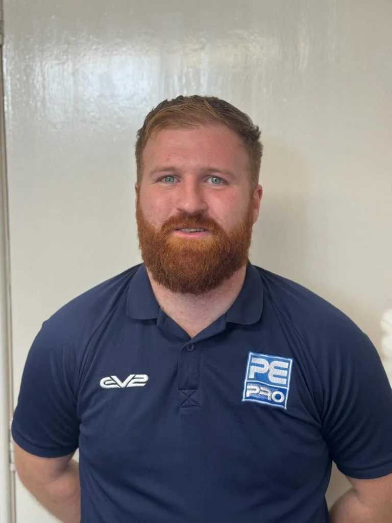 A man with a red beard and short light brown hair, wearing a navy blue polo shirt with logos, standing against a plain off-white wall.