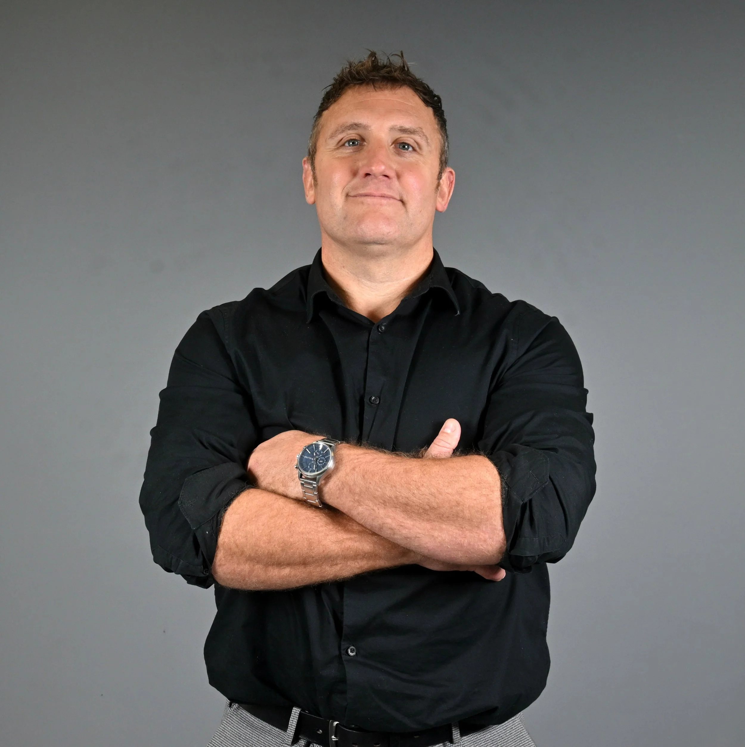 A man with short, messy brown hair and blue eyes, wearing a navy blue collared shirt with the letters 'PE' on it, standing against a plain white wall.
