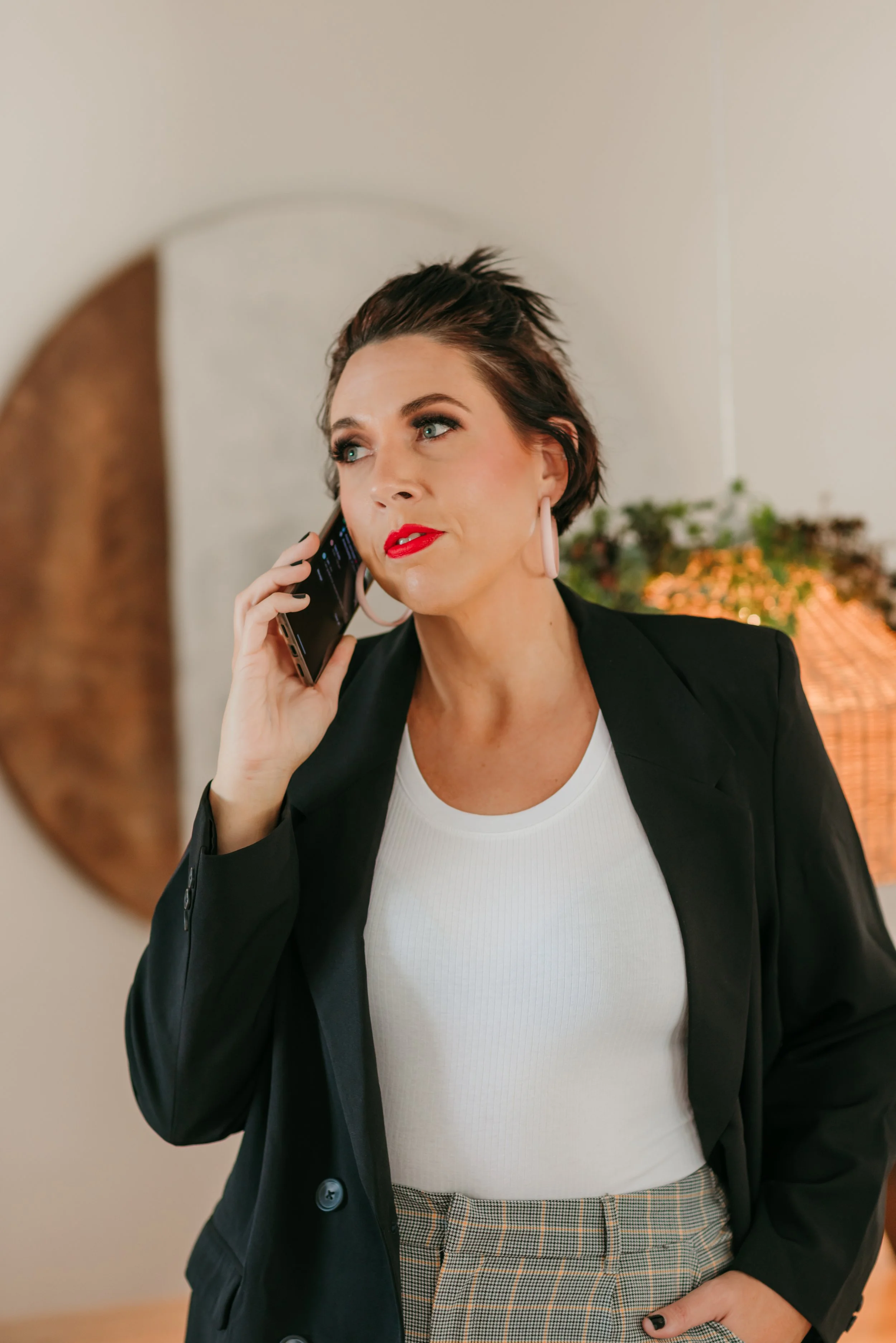 A woman with short dark hair and bright red lipstick talking on her smartphone, wearing a black blazer, white top, and plaid pants, standing indoors with a round mirror and a plant in the background.
