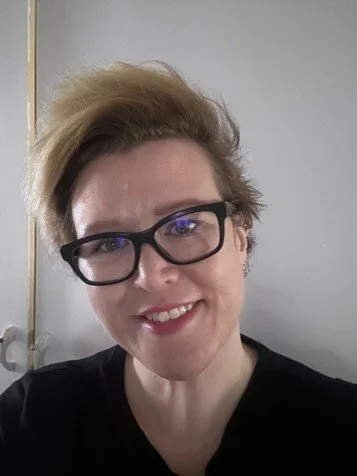 A woman with short blonde hair, black glasses, and a black shirt smiling in front of a gray background.