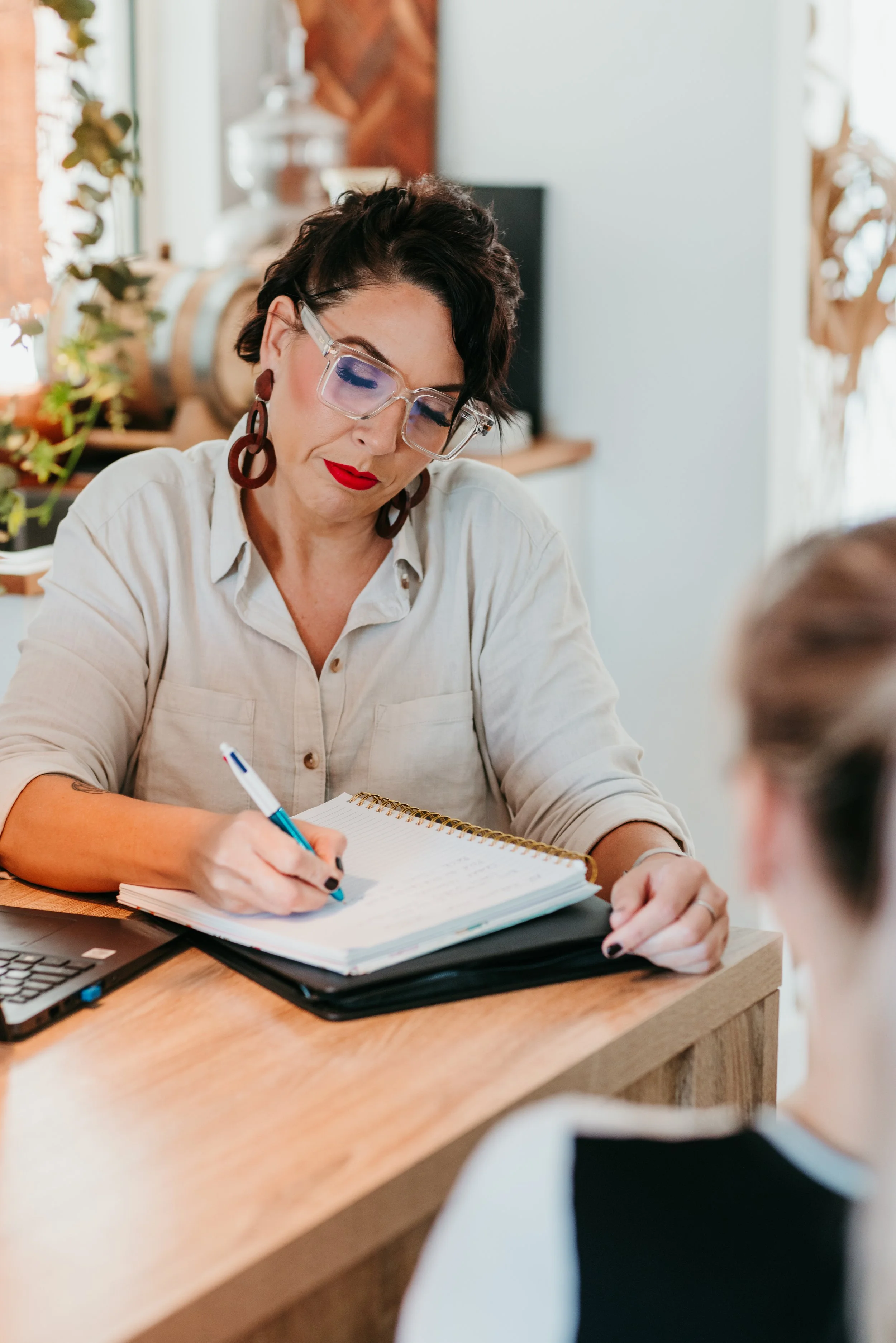 A woman with short dark hair, large glasses, and bold red lipstick writing in a notebook at a wooden table during a meeting or interview.