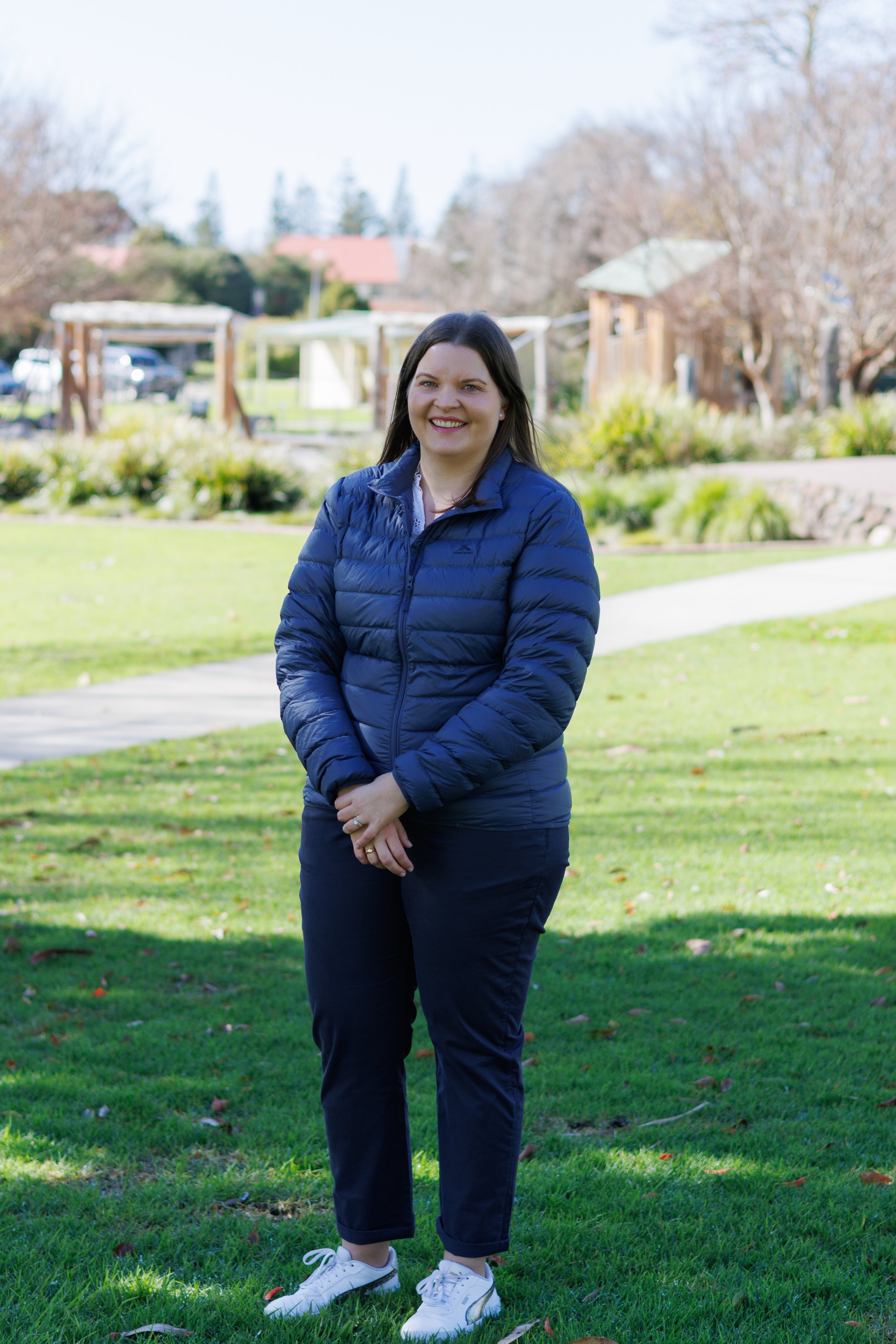 Bethany Findlay, 2025 City of Albany Council Candidate standing on a grassy park in daylight, wearing a blue puffer jacket, dark pants, and white sneakers, smiling at the camera with trees and a building in the background. Election
