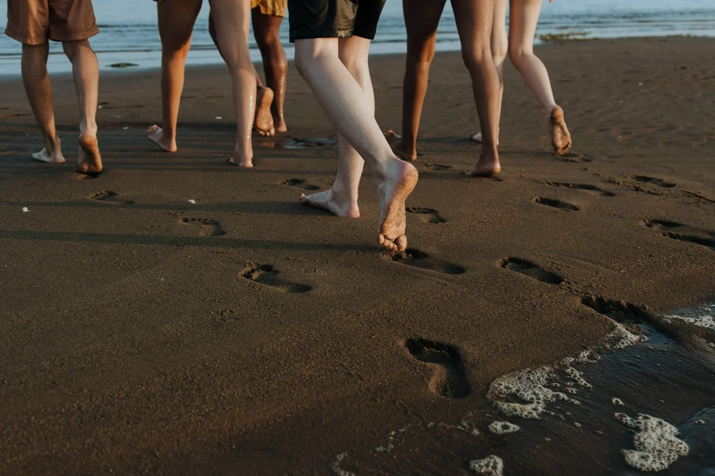 People walking on a sandy beach near the ocean at sunset, leaving footprints in the wet sand.