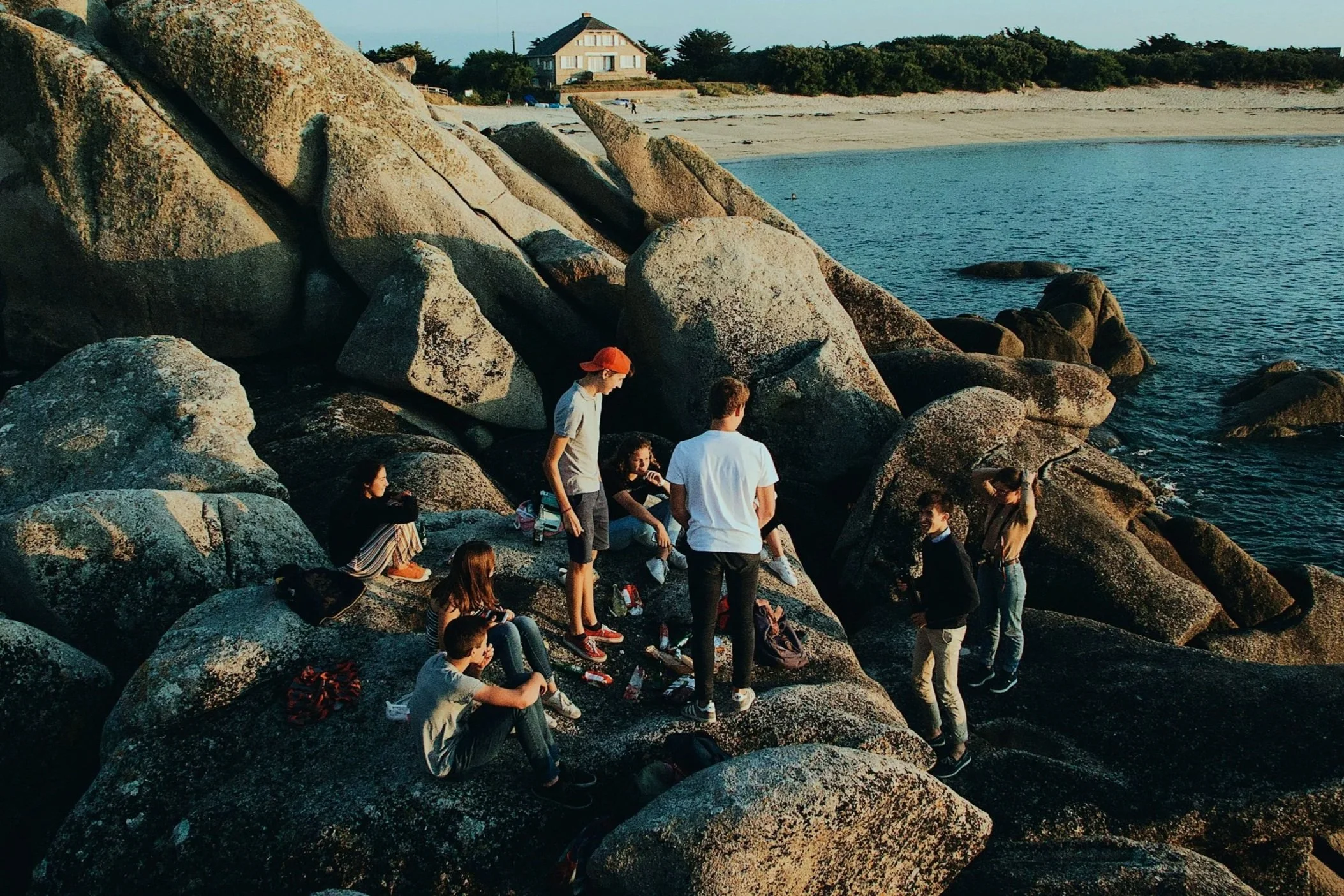 Group of people gathered on large rocks by the shoreline at sunset, with a house and trees in the background, near a beach and water.