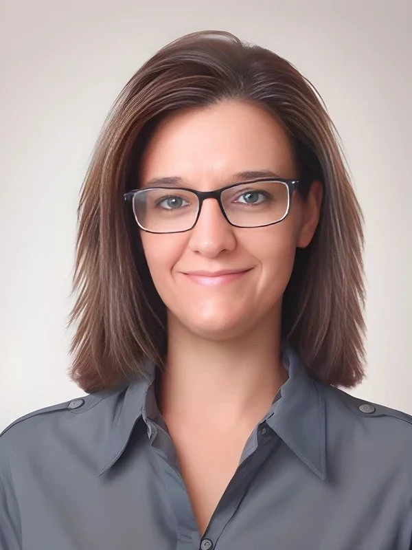 A woman with shoulder-length brown hair wearing glasses and a gray button-up shirt, smiling in a close-up portrait against a neutral background.