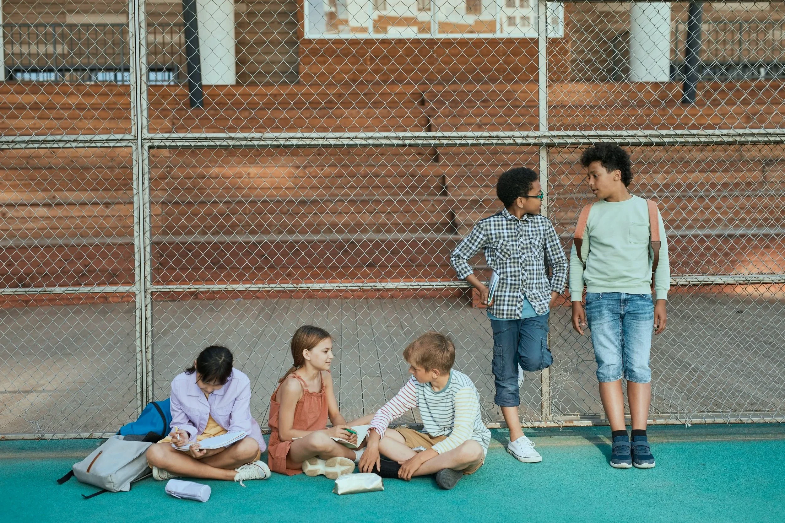 Five kids, three boys and two girls, waiting outside a fenced area, some sitting on the ground and two standing, with backpacks, on a green surface