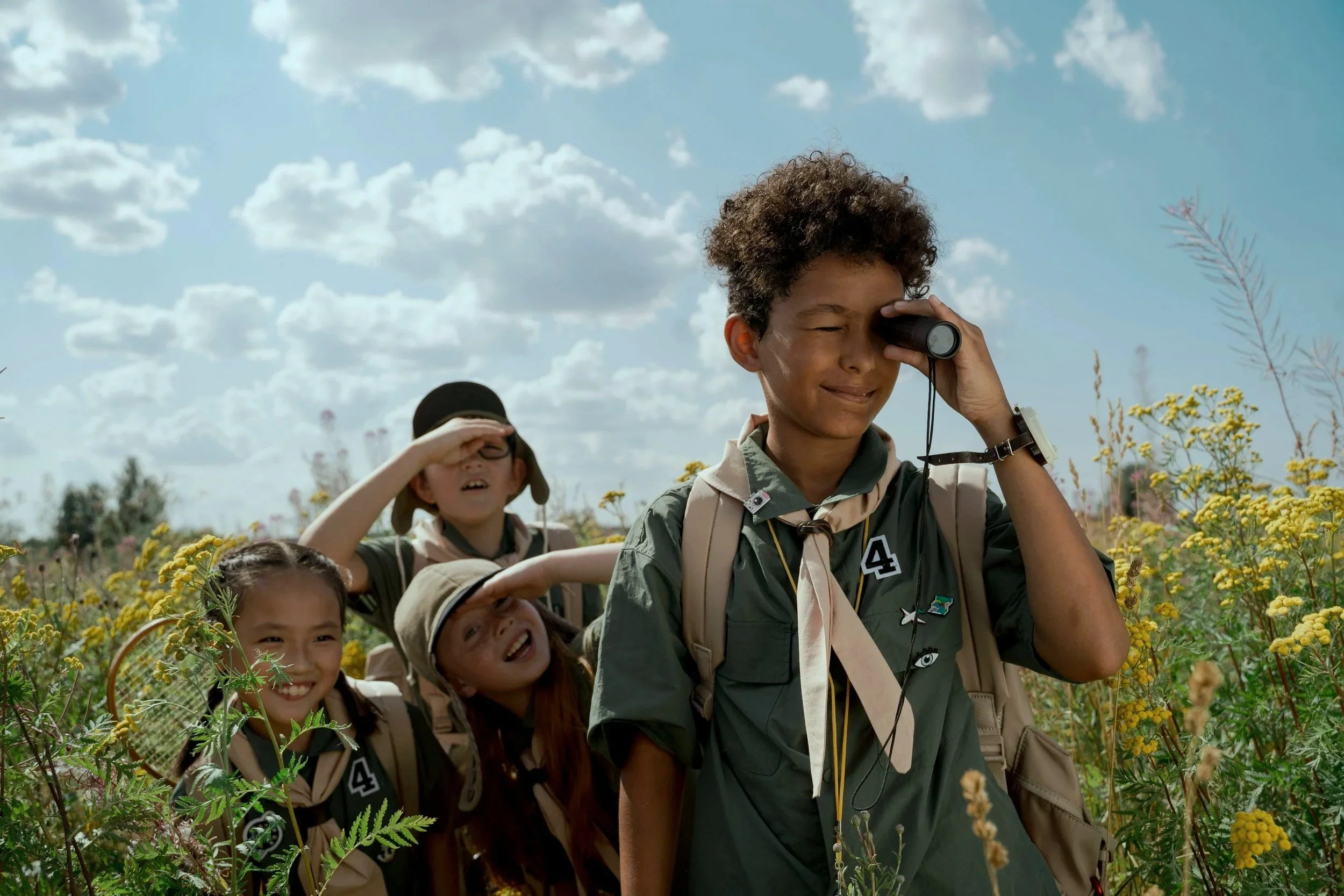 Group of young scouts exploring outdoors, looking through binoculars and shielding their eyes from the sun in a field of wildflowers under a blue sky with clouds.