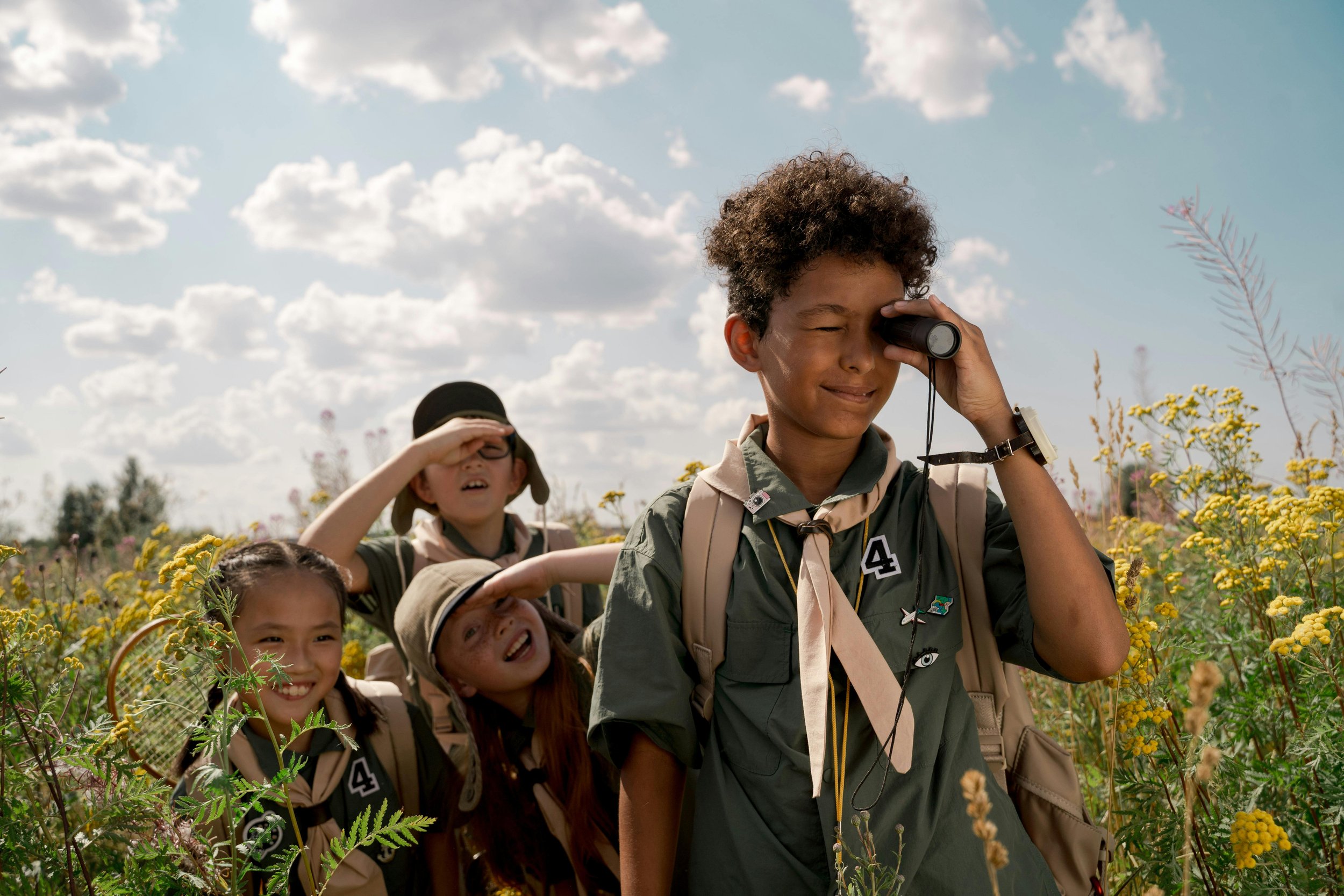 Four young scouts hiking through a field of yellow wildflowers under a partly cloudy sky, using binoculars and sun hats.
