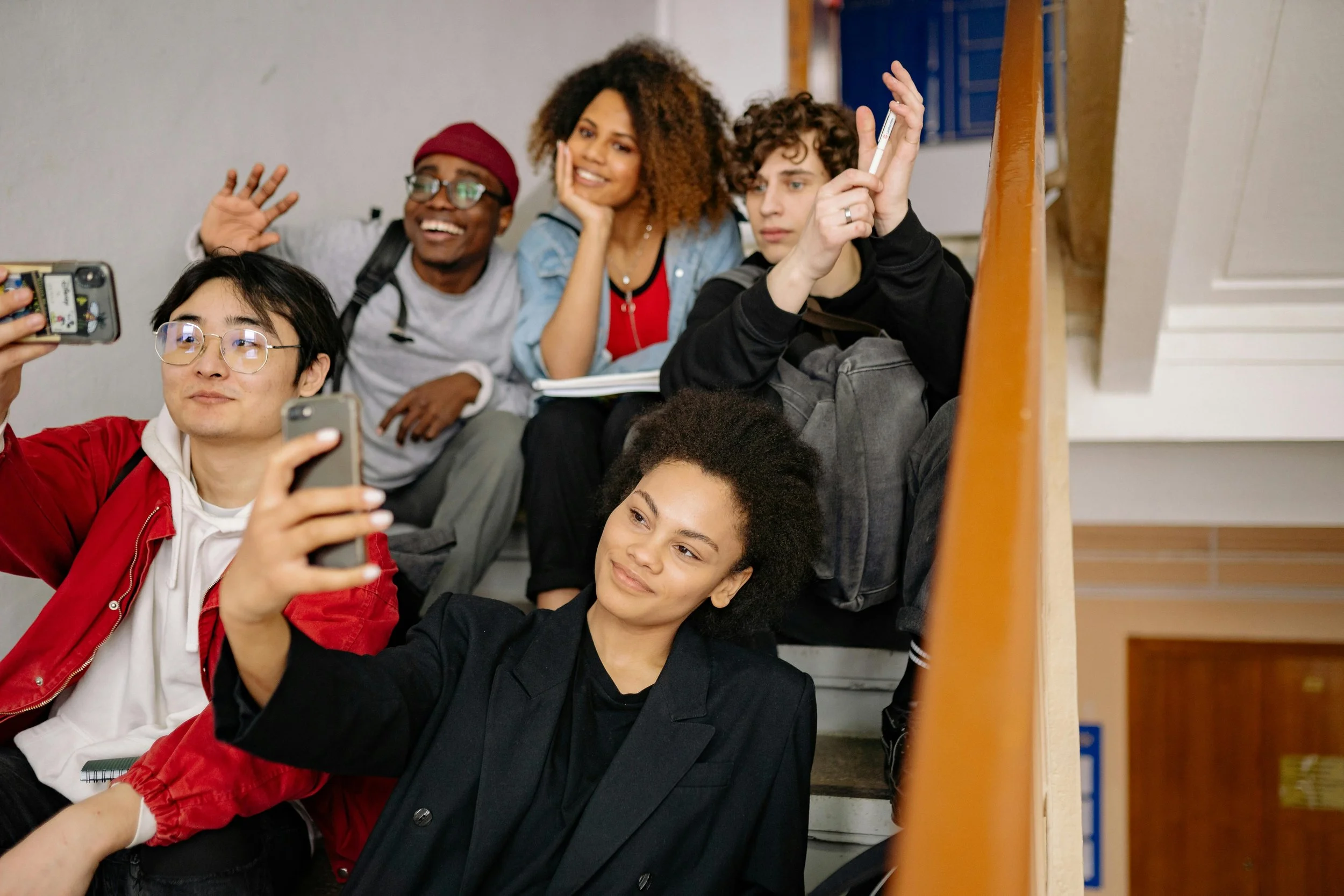 Group of five diverse young students sitting on a staircase, taking selfies and smiling.
