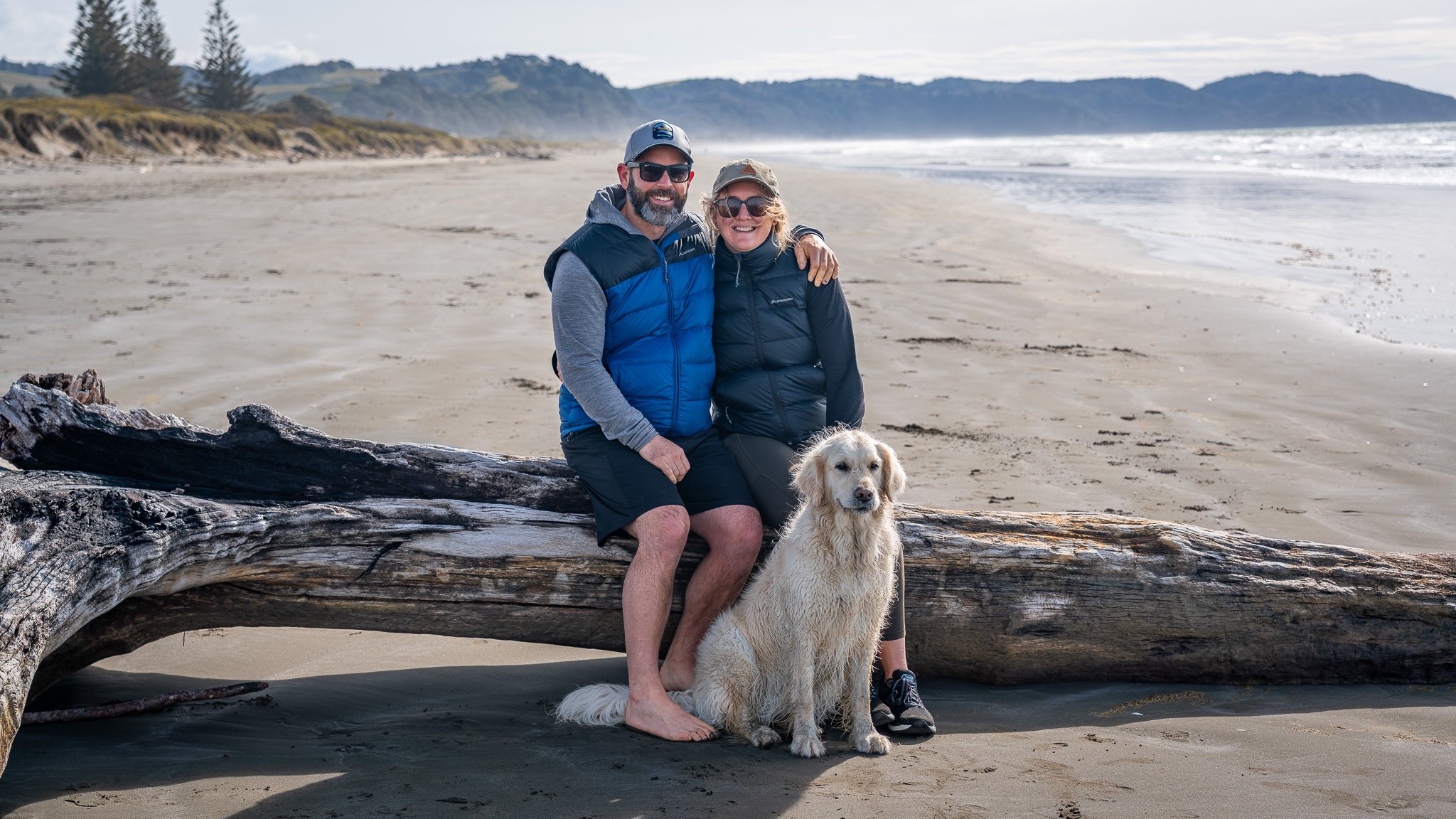 A smiling couple and their dog at the beach, sitting on a large driftwood log with ocean waves and distant hills in the background.