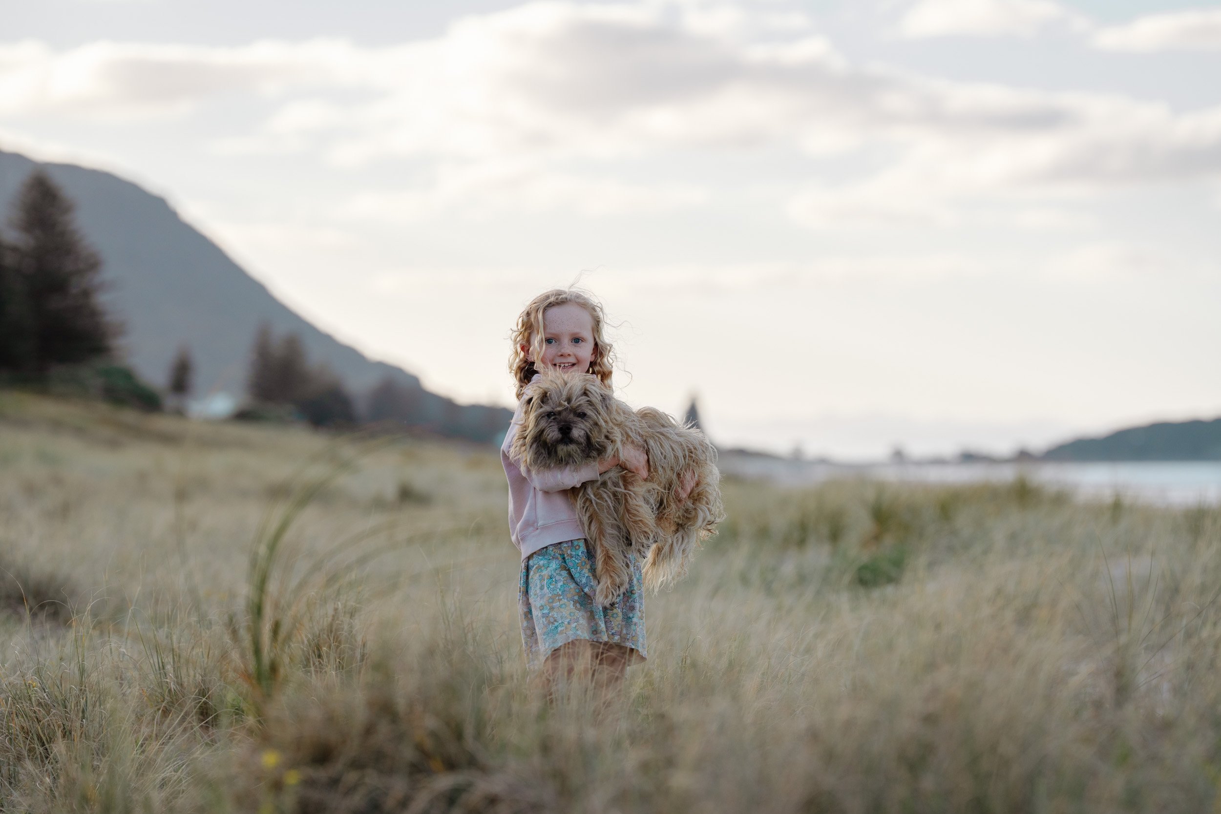 A young girl with curly blonde hair wearing a pink jacket and patterned shorts holding a shaggy brown dog in a grassy field near a body of water with mountains in the background.