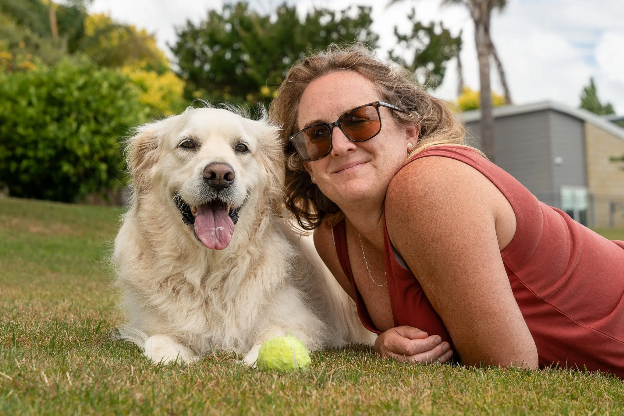 A woman lying on grass next to a golden retriever dog with a tennis ball, smiling in a park with trees and a house in the background.