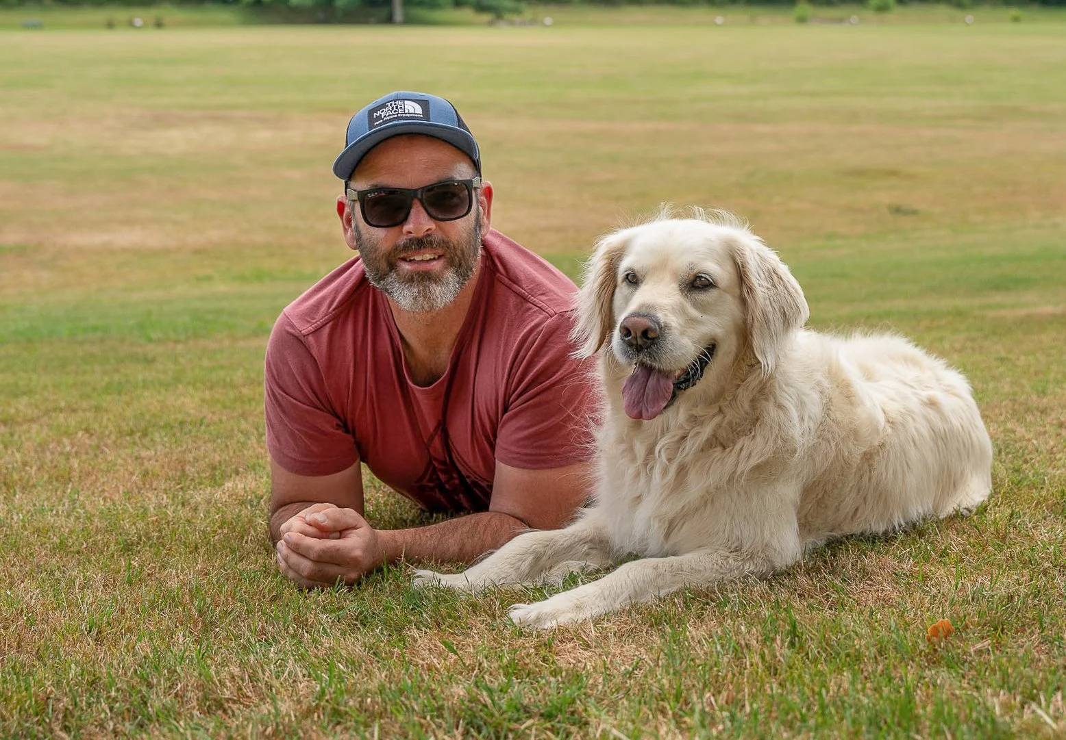 A man lying on the grass next to his golden retriever dog on a large open field.