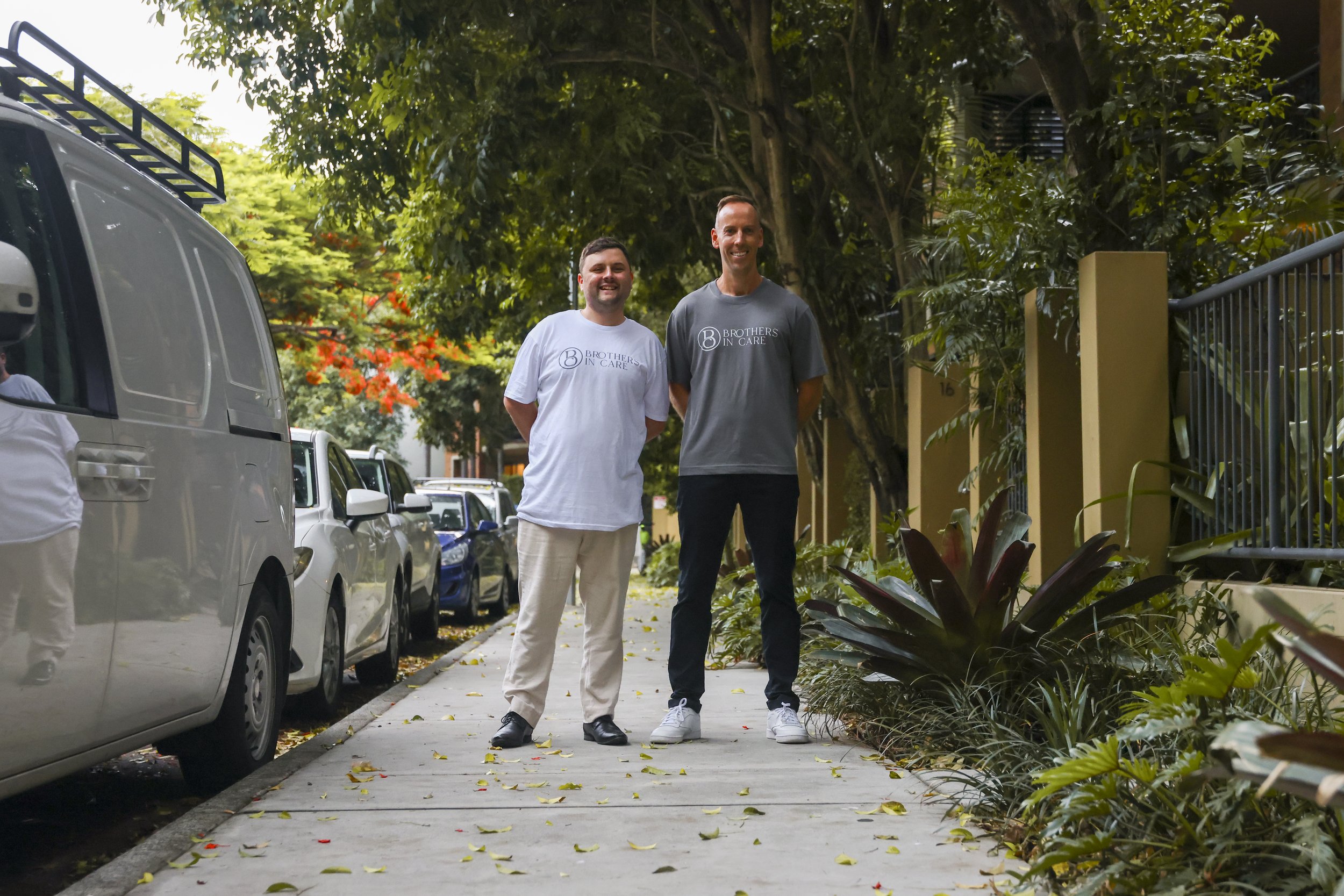 Two smiling men standing on a sidewalk with parked cars and lush greenery in the background.