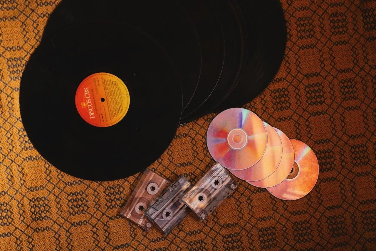 A collection of old black vinyl records, some CDs, and cassette tapes on an orange patterned fabric surface.