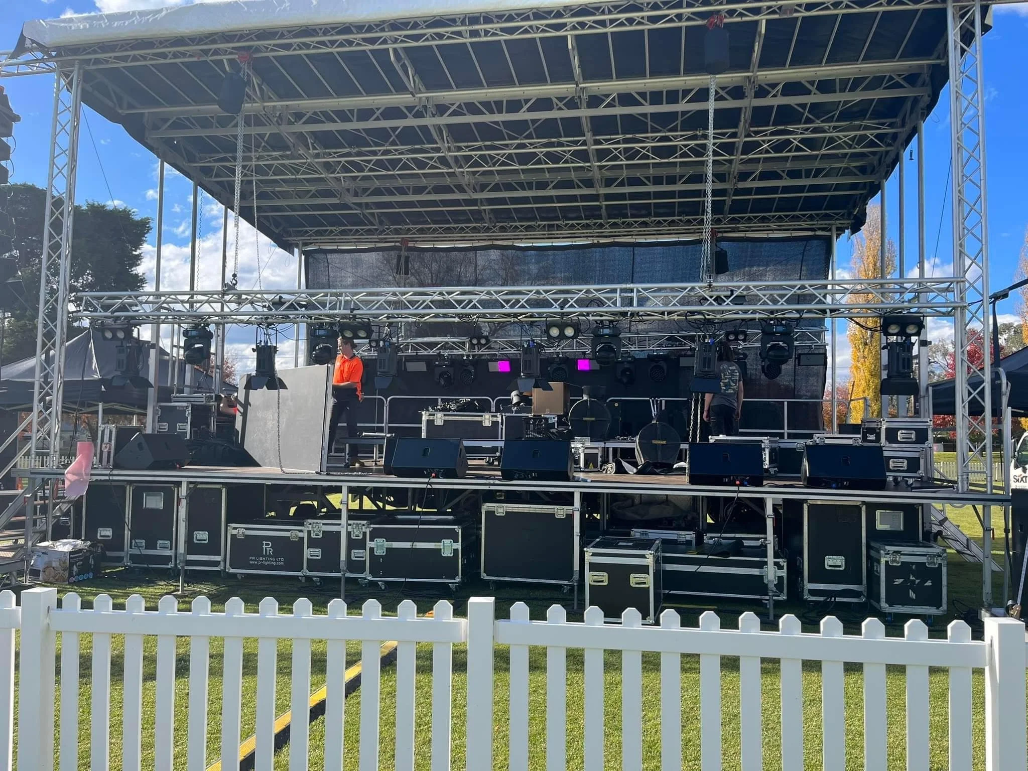 Empty outdoor stage with lighting and sound equipment, two crew members working, surrounded by a white fence, during daytime with partly cloudy sky and trees in the background.