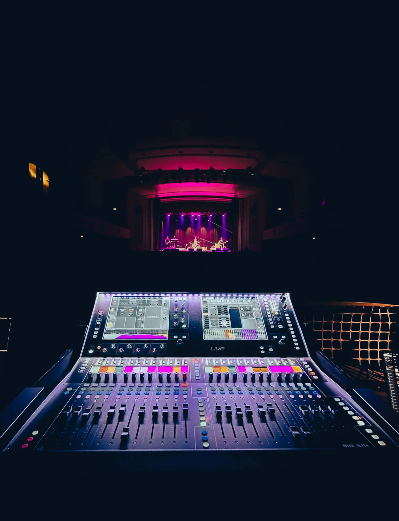 A professional audio mixing console in a concert hall, with a stage performing live music in the background under pink and purple lighting.