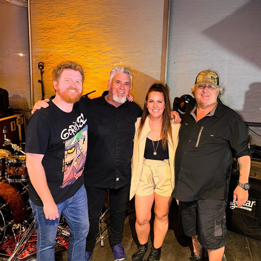 The members of the band Savage Groove standing together and smiling, in an indoor setting with musical equipment in the background.