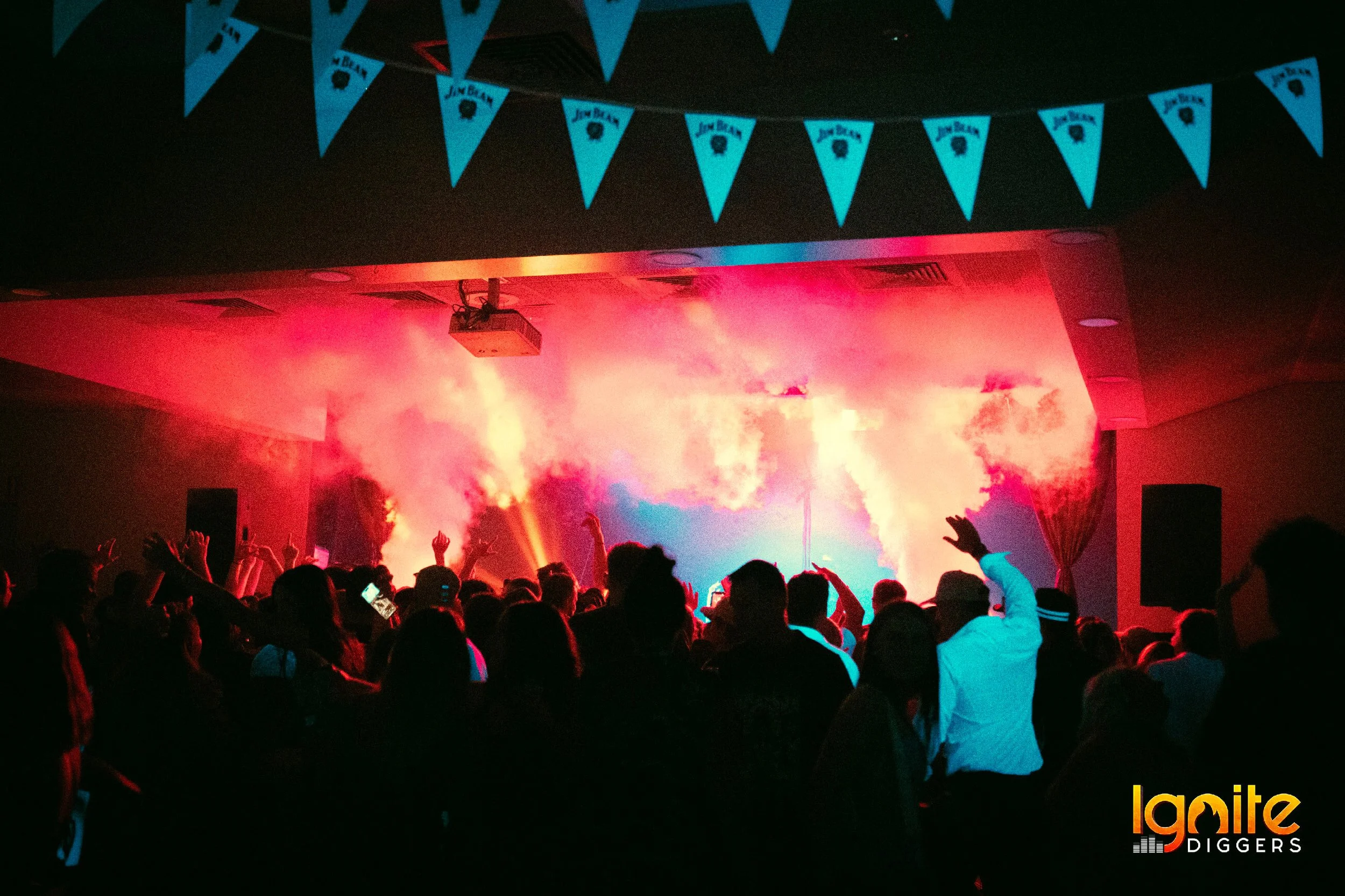 Crowd at a concert with smoke and colorful lights on stage, blue pennant flags hanging above, Ignite Diggers logo in the bottom right corner.