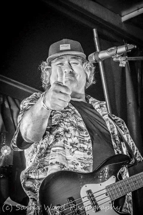 Fat Man Solo holding a guitar and pointing at the camera with a thumbs-up gesture on stage with a microphone.
