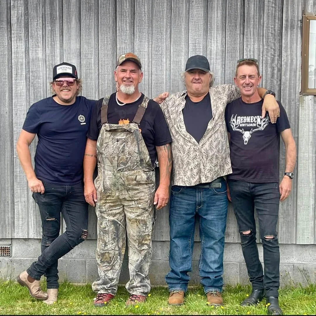 The members of the band Redneck Gentlemen standing together in front of a wooden wall, smiling and posing for the photo, with their arms around each other.
