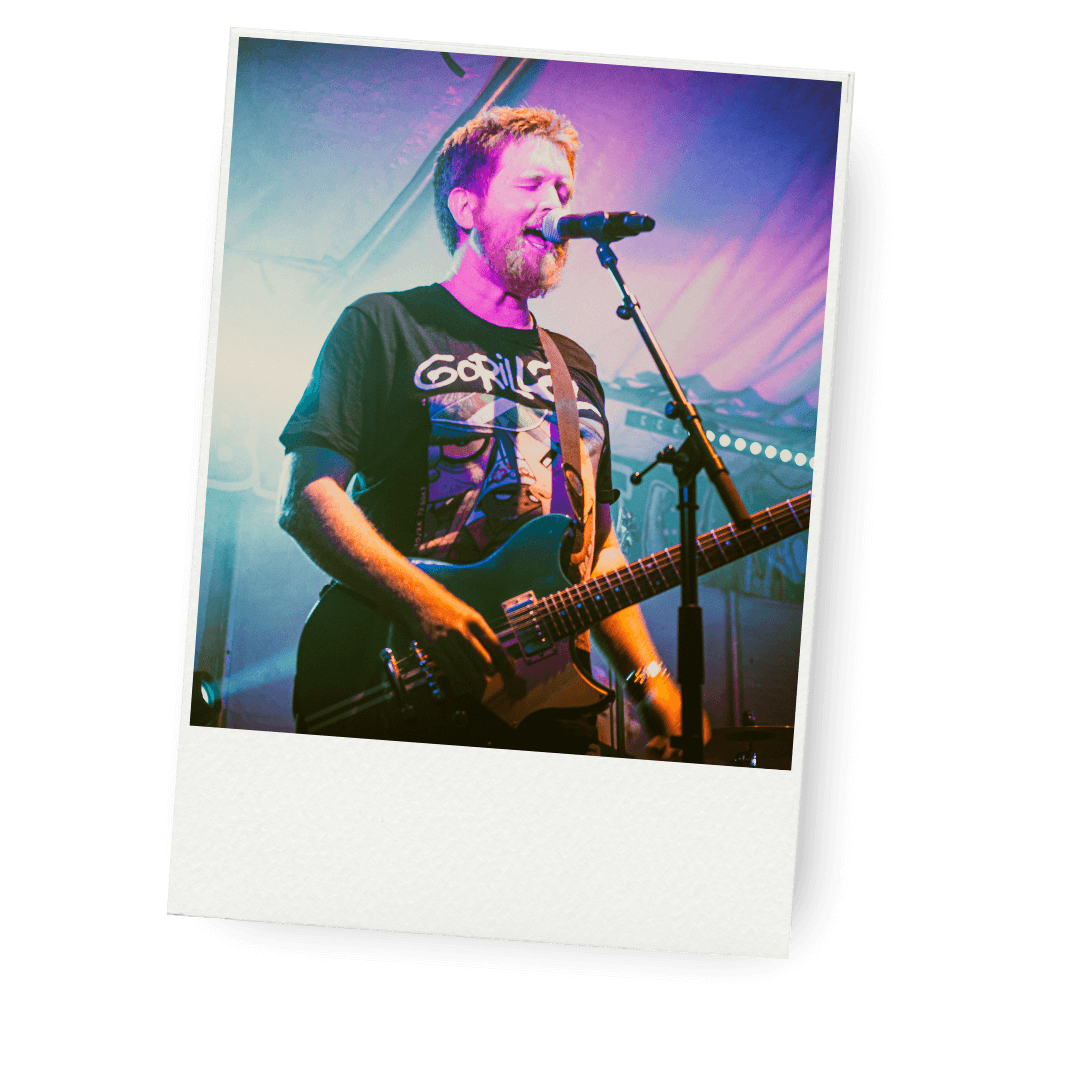 Polaroid picture of a member of Savage Groove with a guitar singing into a microphone on stage, wearing a black t-shirt with a graphic design and a 'Gorillaz' logo, with colorful stage lighting.