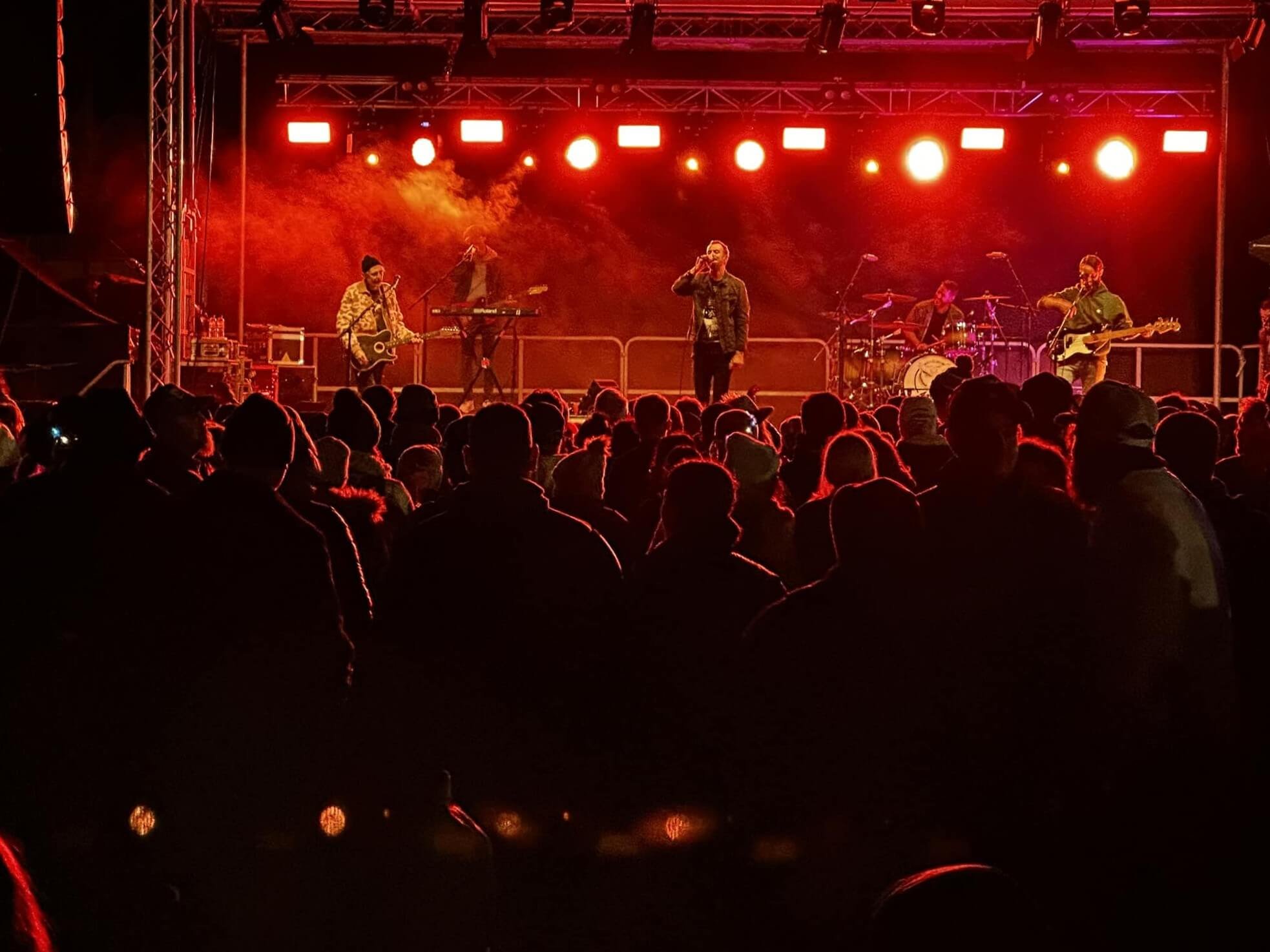 A band performs on stage at night with red and orange lighting, with an audience watching in the foreground.