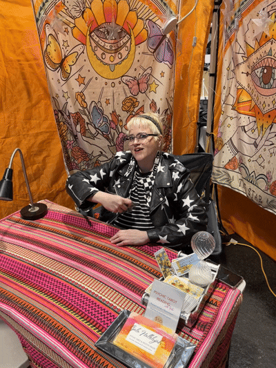 A woman with glasses and short blond hair sitting at a table with a colorful striped tablecloth, in front of a backdrop with psychedelic and nature-themed artwork featuring butterflies, eyes, and a sun with a face. She is wearing a leather jacket with star patterns.