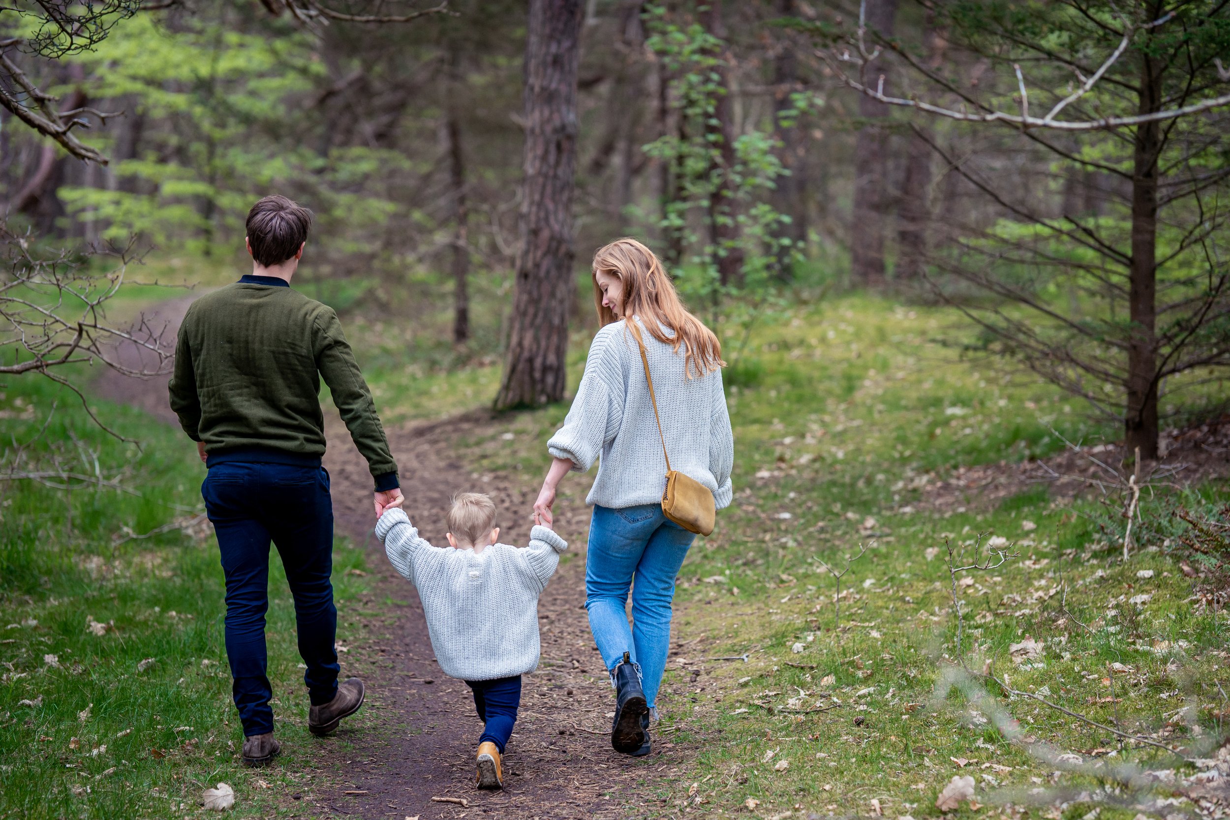 En familie går tur i skoven med to voksne og to børn, set fra bagfra, med grønne træer og stien foran dem.