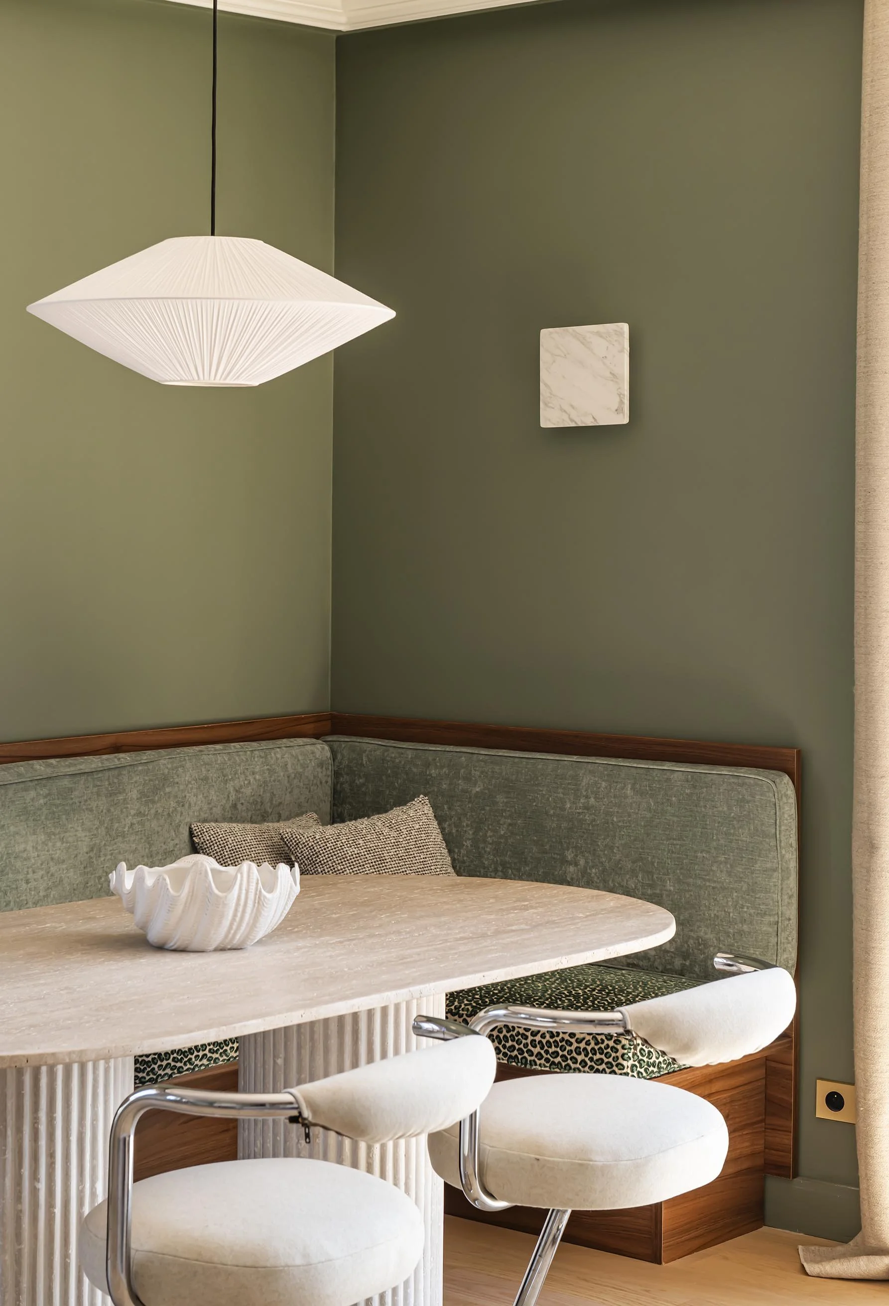 Coin repas avec une table en marbre, deux chaises blanches et un banc vert avec des coussins, mur vert avec un cadre en marbre, luminaire blanc suspendu, rideau beige à droite.