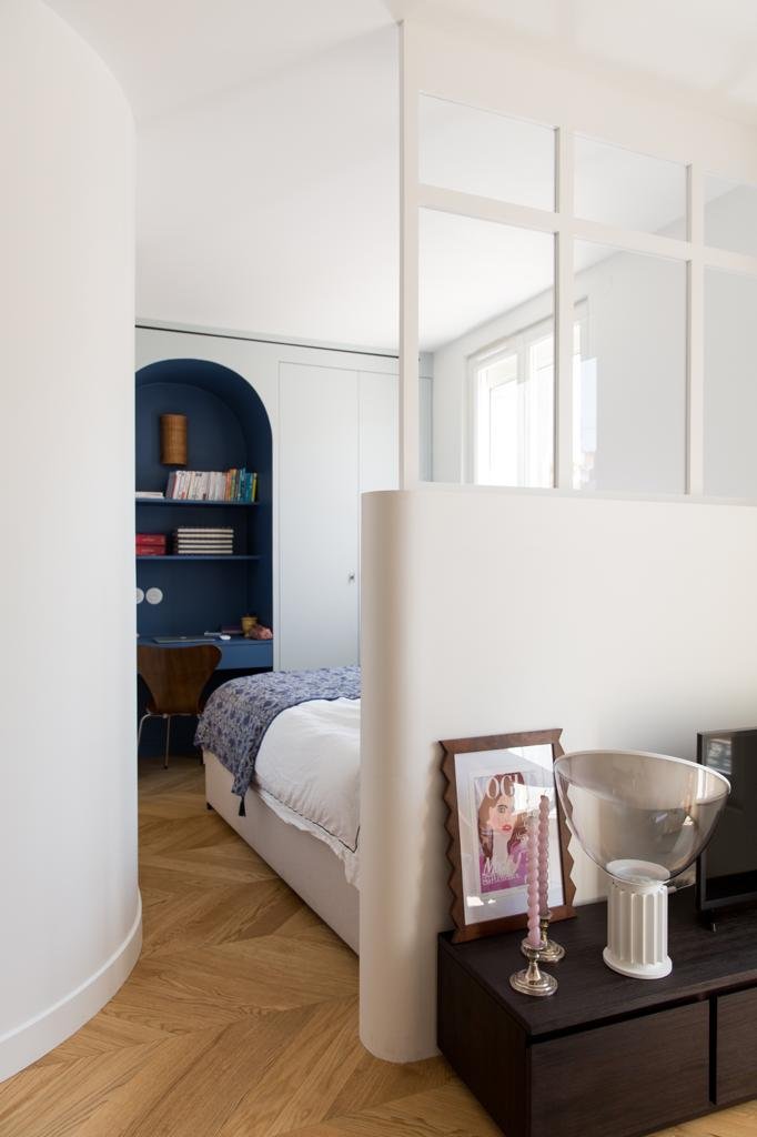 Intérieur d'une chambre moderne avec un mur blanc, un lit avec une couverture grise, une étagère bleue encastrée avec des livres et une lampe, et une console avec une photo encadrée, une bougie et un vase transparent.