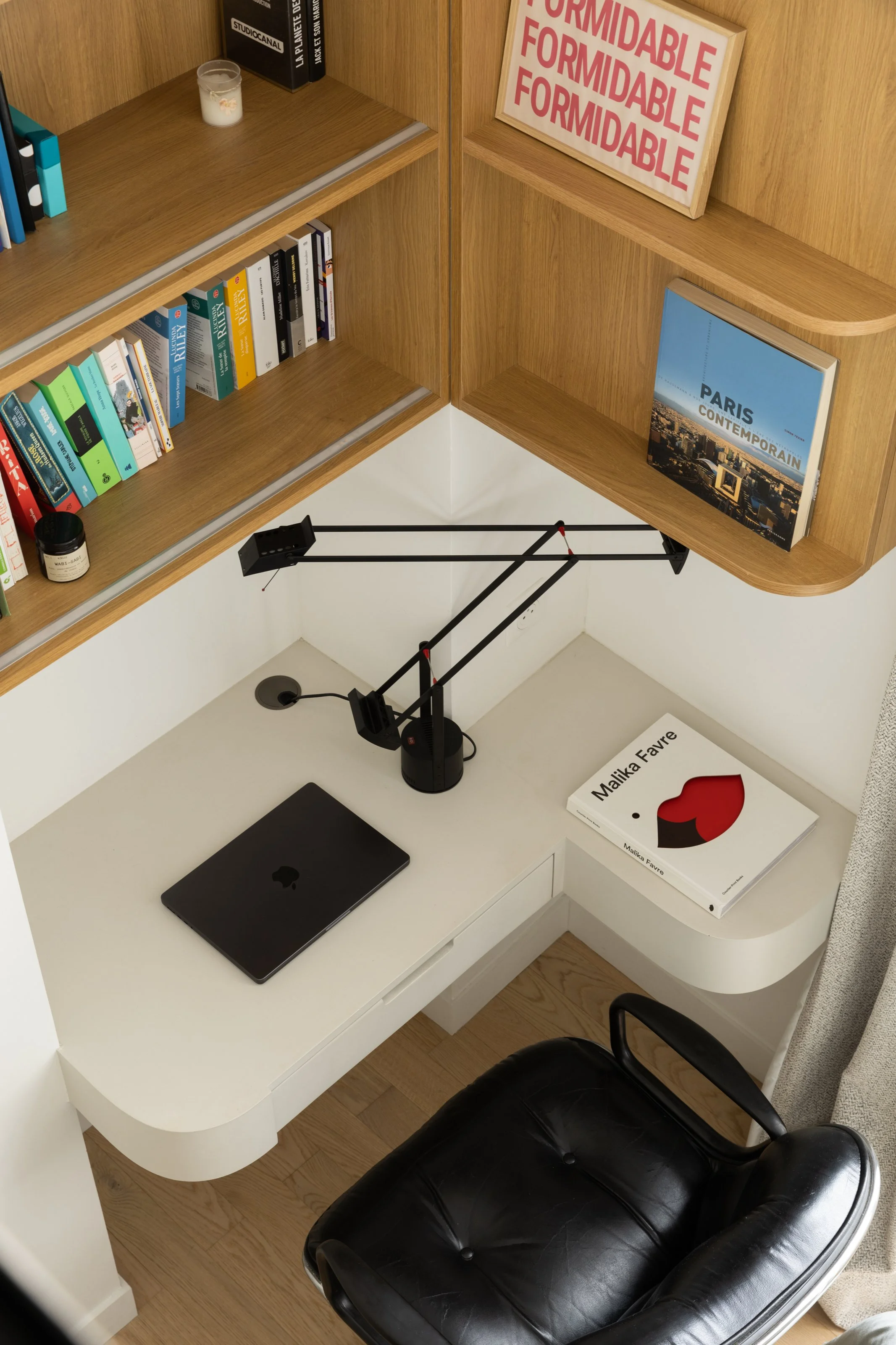 Un coin bureau avec un bureau blanc, un ordinateur portable noir, un livre blanc intitulé "Malika Favre" avec un design rouge et noir, une lampe de bureau noire, une chaise en cuir noir, des étagères en bois contenant des livres et une affiche "Formidable" en cadre. Il y a aussi une affiche "Paris Contemporain" sur l'étagère.