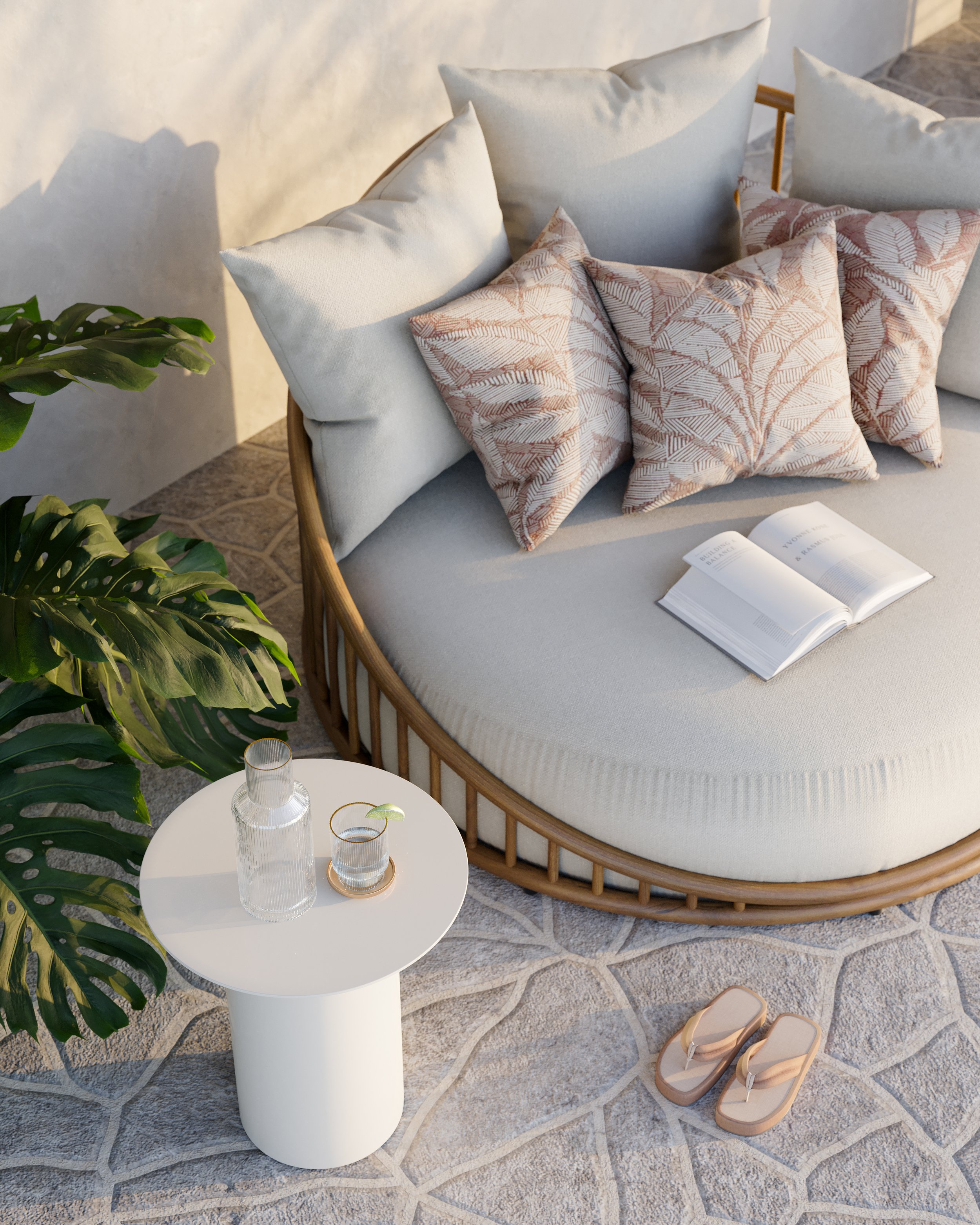 A light-colored, curved sofa with multiple pillows, including patterned and plain ones, next to a white round side table. A bottle and a glass with a slice of lime are on the table, and tan sandals are on the floor. There is a large green plant to th