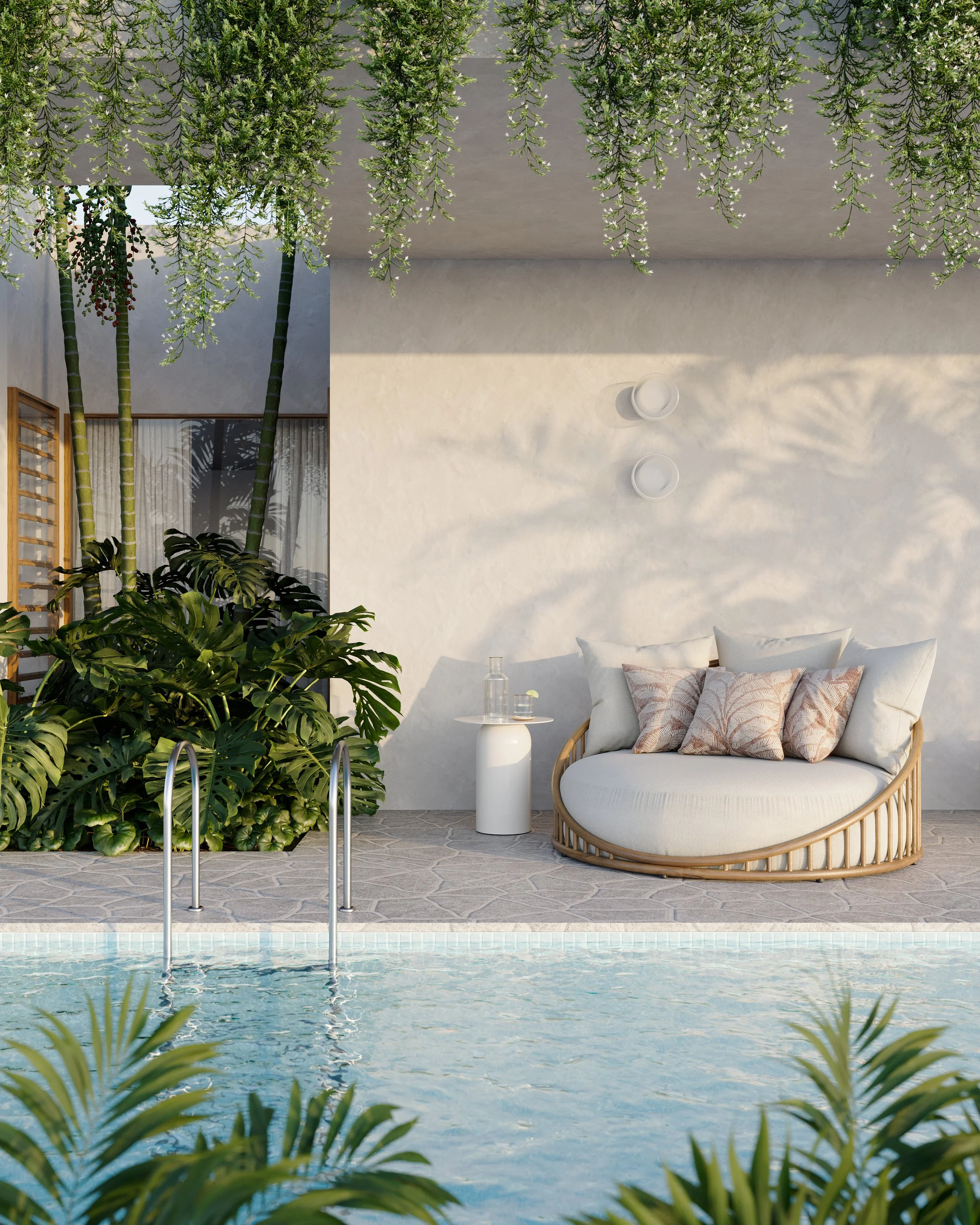 A cozy outdoor seating area with a round wicker couch with white cushions and decorative pillows, a small white side table with a water bottle and glass, lush green tropical plants, hanging greenery overhead, and a pool in the foreground.