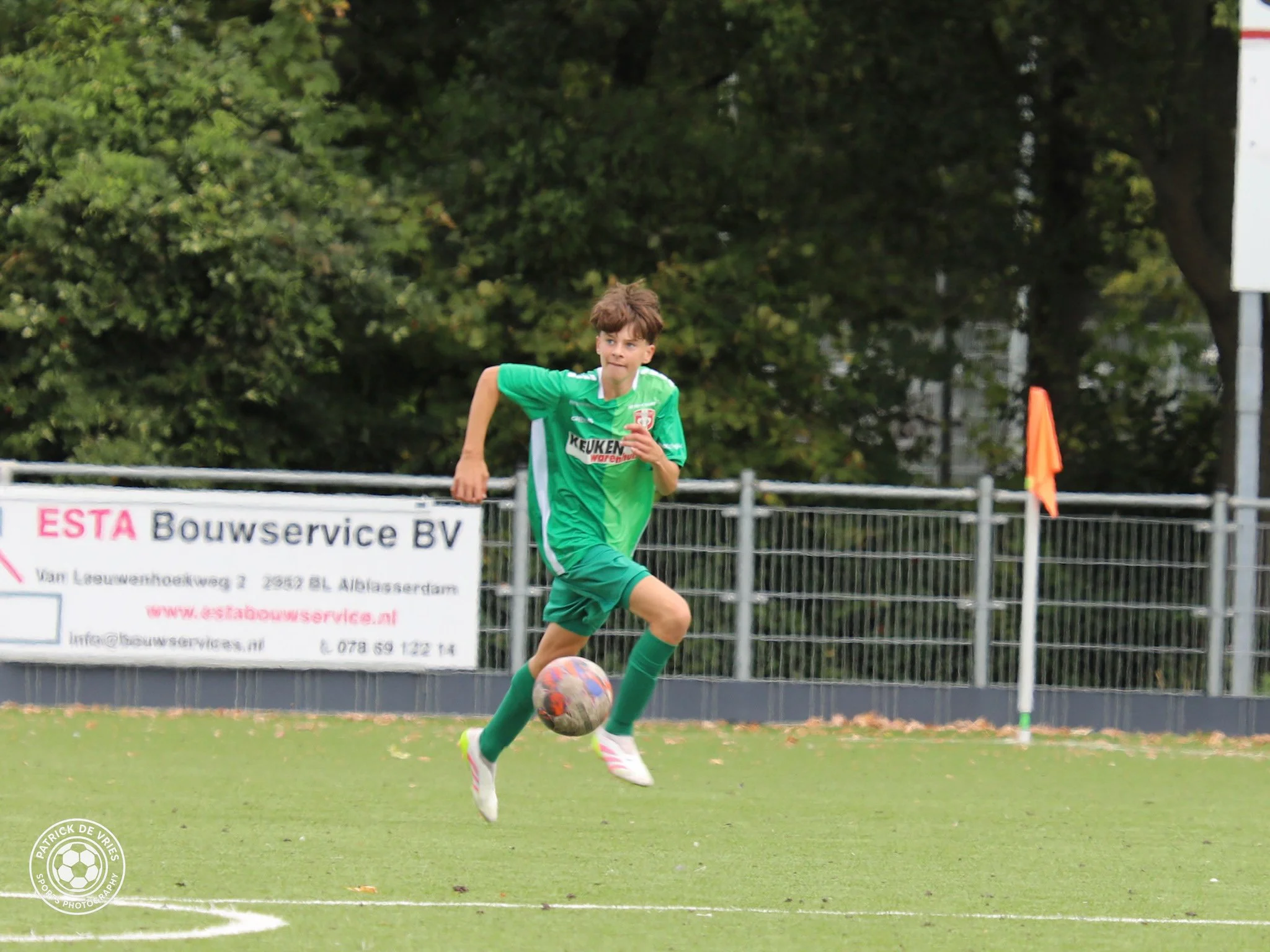 Jongen die een voetbal trapt op een voetbalveld, met bomen op de achtergrond en een reclamebord.